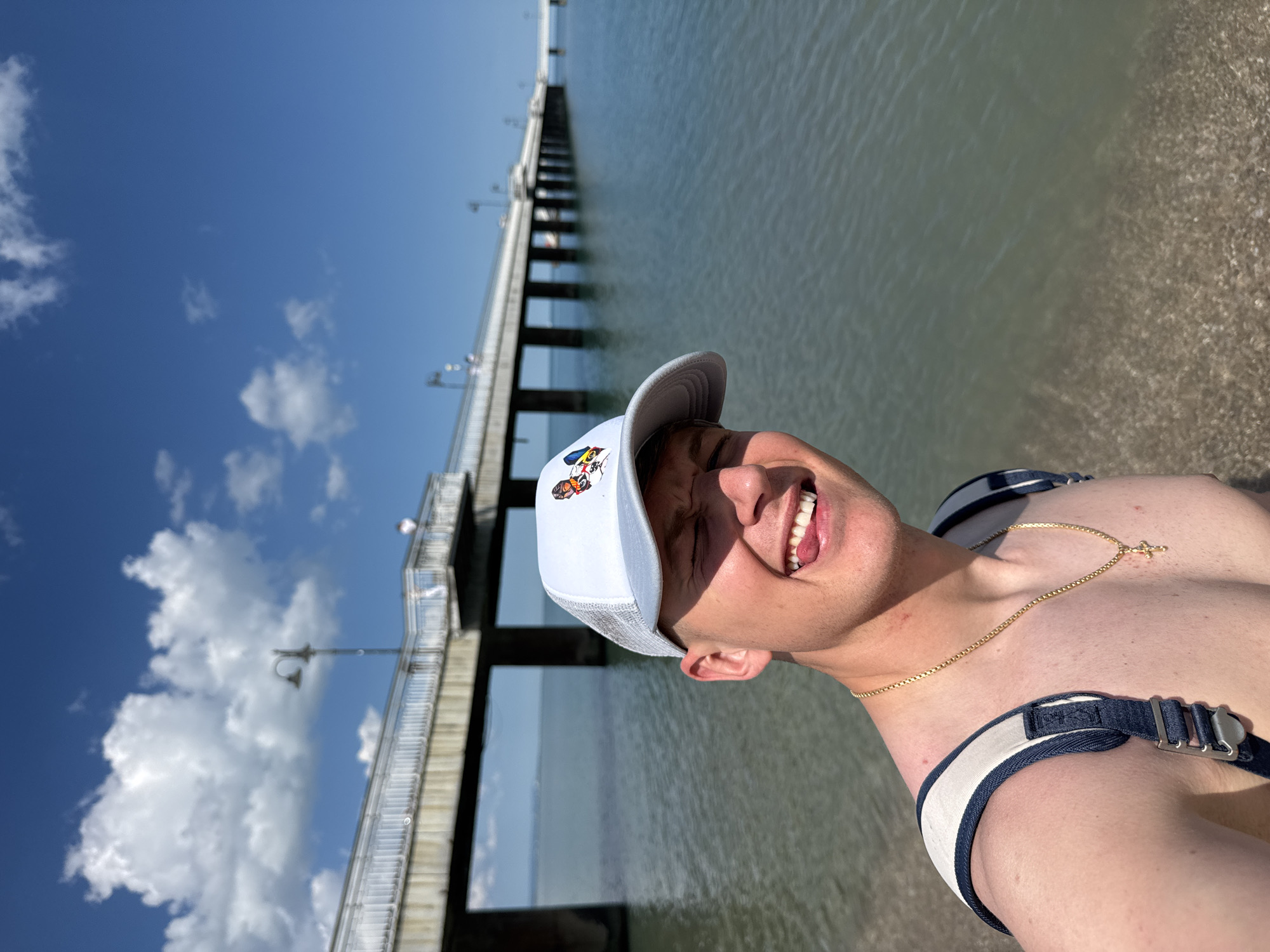 A close-up portrait shows a man smiling at the camera, wearing a white baseball cap with a cartoon design, a gold necklace with a cross pendant, and a harness-like garment. He is positioned near the shoreline, with water and a sandy beach visible to the right. In the background, a long pier stretches out over the water. The sky is blue with scattered clouds. The location is Burgas, Bulgaria, as evidenced by the Burgas Pier, a recognizable landmark in the city.