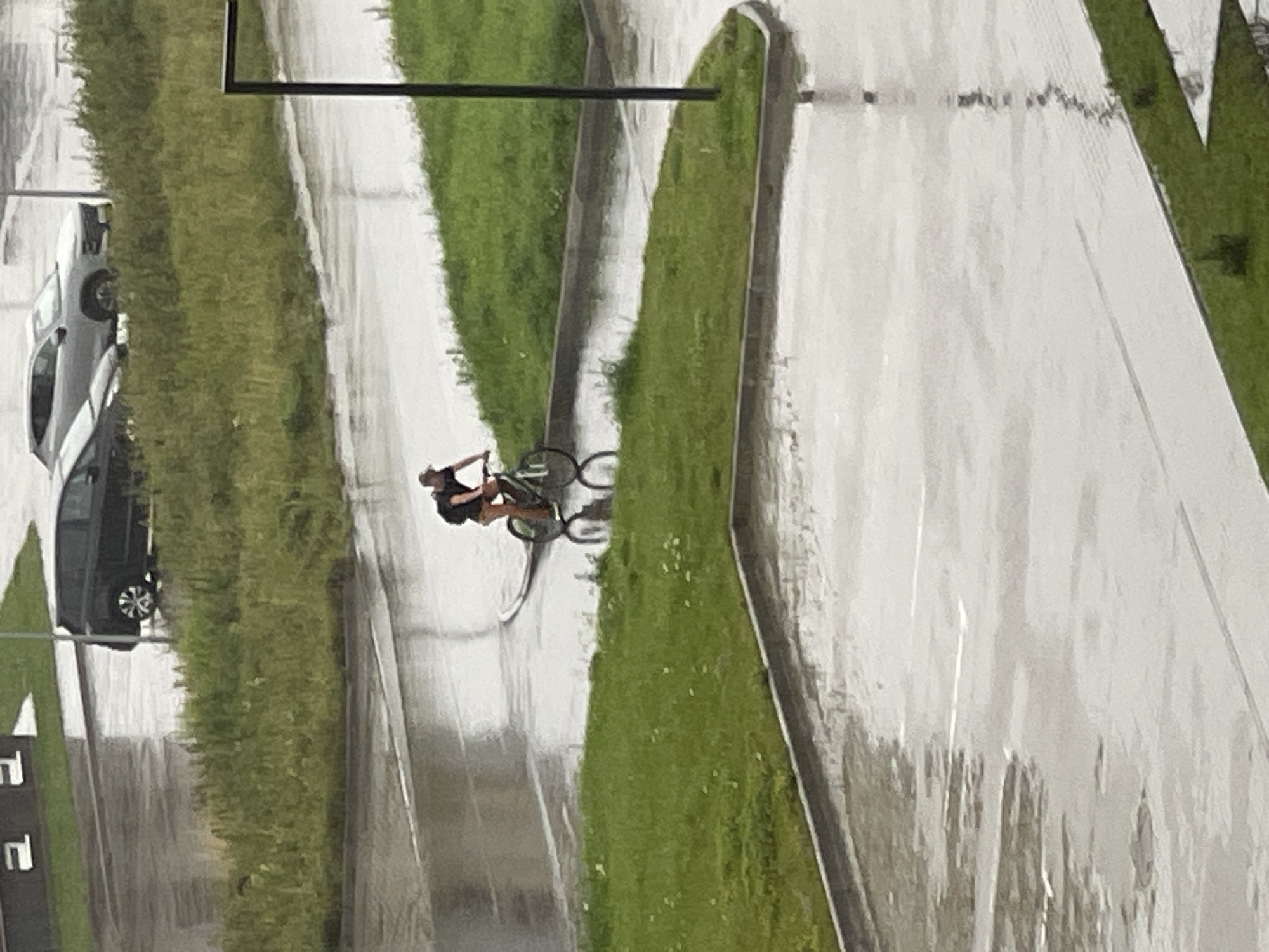 A person is cycling on a wet paved surface in Zelenogradsk, Russia. The person is wearing a dark t-shirt and shorts. The bicycle is dark colored. There is green grass to the left and right of the paved surface. In the background, there are cars parked on a wet road. The overall environment appears to be a residential or urban area experiencing rain.