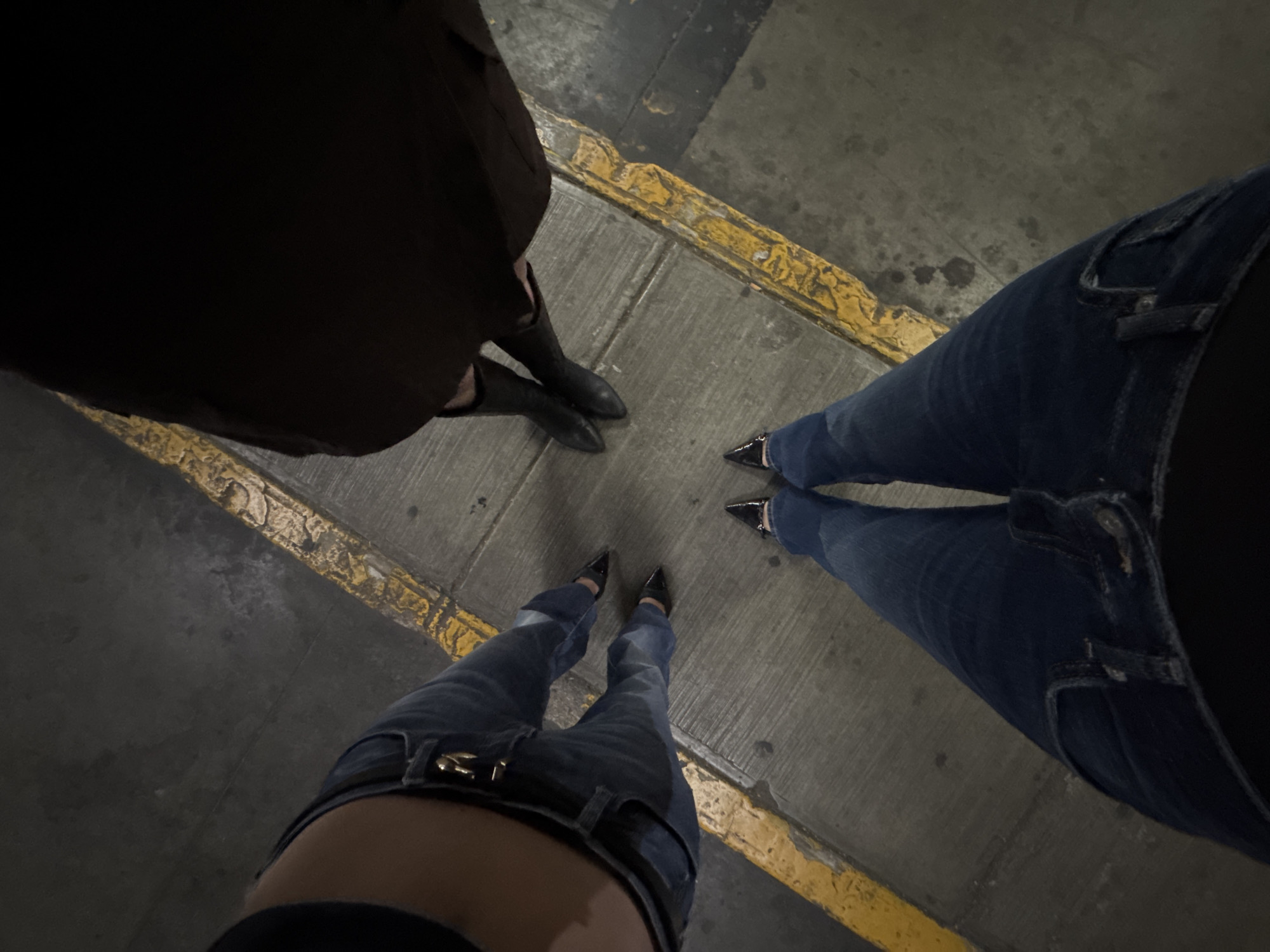 The image shows a downward-angled view of three people standing on a grey concrete surface with yellow painted lines, potentially in Mexico City, Mexico. One person on the left wears dark-colored boots under a dark, seemingly brown, coat. Another person on the right wears jeans and pointed black shoes. The third person, located at the bottom, also wears jeans with a belt and pointed black shoes. All three are positioned in a roughly triangular formation, with their feet close to the center of the frame. The concrete has some minor stains and discoloration. The yellow lines appear aged and worn.