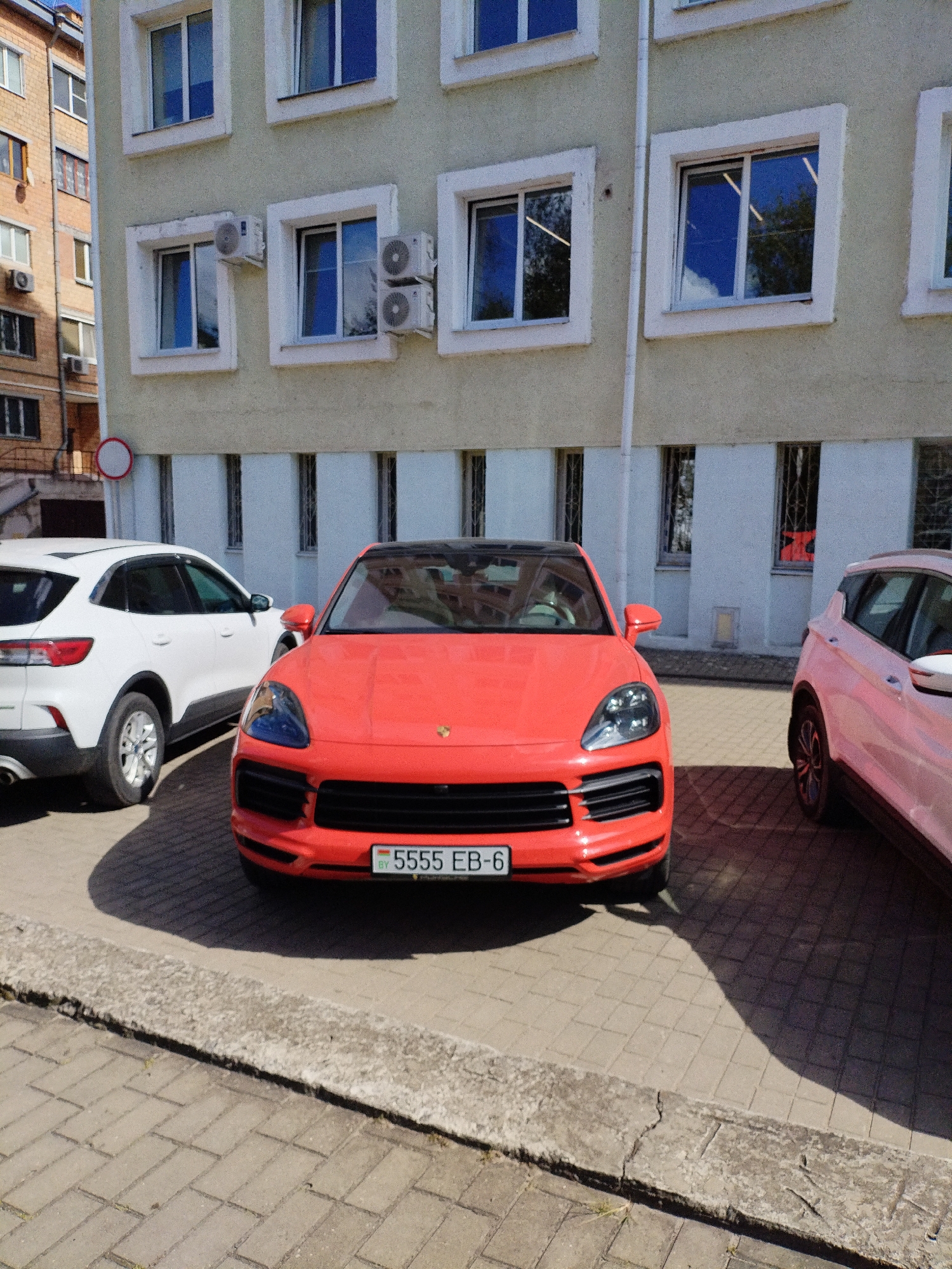 In the city of Mahilyow, Belarus, a bright orange Porsche Cayenne is parked on a paved lot in front of a light-colored building with multiple windows. To the left of the Porsche, a white SUV is partially visible. To the right, the rear of a pink SUV is seen. The building has several windows with air conditioning units mounted below them. A traffic sign with a red circle and a white horizontal bar is visible to the left of the building. The license plate on the Porsche reads "5555 EB-6". The scene appears to be captured during daylight hours under a clear sky.