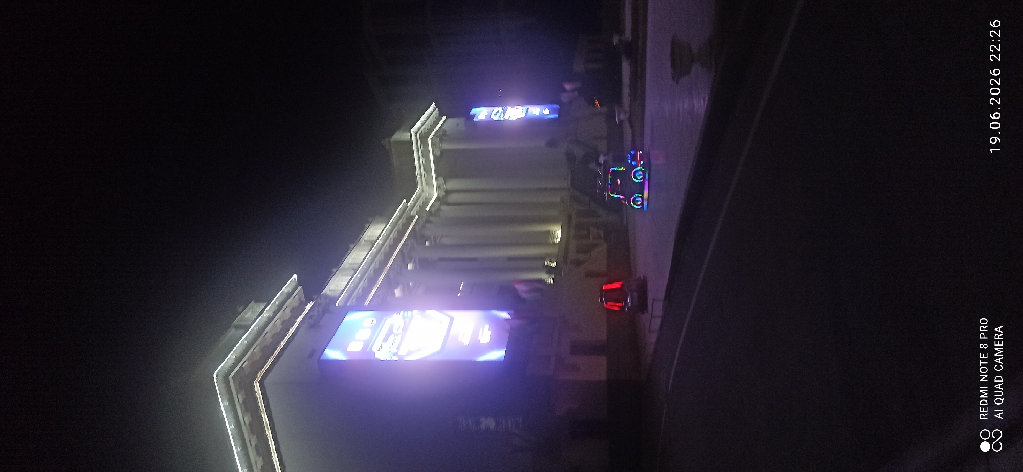 The image is an outdoor nighttime shot of a grand building, likely a museum or government building, with classical architecture featuring columns and ornate trim illuminated by white lights. A large, vertical digital billboard displaying colorful graphics is attached to the left side of the building. Two small, brightly lit vehicles resembling bumper cars are positioned on the paved area in front of the building, one to the left with red lights and another to the right with multi-colored lights. The sky is dark and hazy. The bottom of the image shows a street in the foreground and text overlays indicating "REDMI NOTE 8 PRO AI QUAD CAMERA" and a date and time stamp "19.06.2026 22:26".