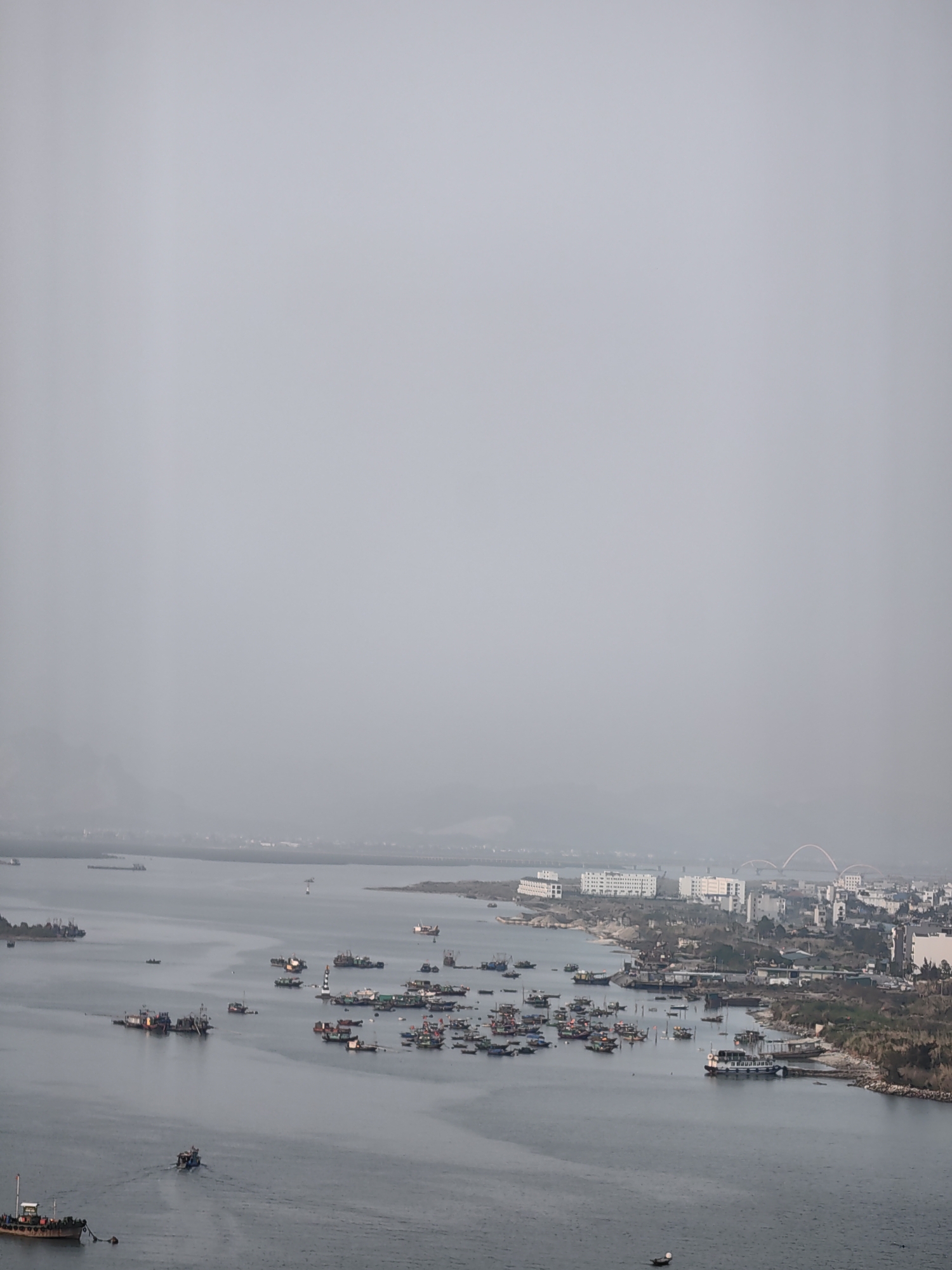 The scene is a view of Ha Long Bay from an elevated position in Hong Gai Ward, Vietnam. In the foreground, the bay is populated with numerous boats of various sizes, mostly fishing vessels. The water is calm with visible wakes from the boats. The middle ground features a peninsula with low-rise buildings and some vegetation. In the background, buildings and a bridge are visible, though they are obscured by a hazy atmosphere. The sky is overcast and uniformly gray, indicative of pollution or fog. The overall scene is industrial with signs of maritime activity and urban development. No people are discernible in the shot.