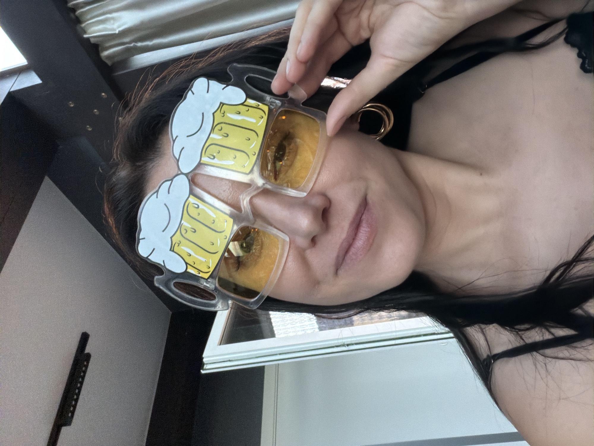 A woman is taking a selfie indoors, wearing novelty sunglasses shaped like beer mugs. She has long, dark hair and is wearing a black camisole. She is holding one of the sunglasses with her right hand. The background includes a ceiling with a light fixture, a window or door, and curtains. The location is not specified as Karsha, Russia; this information appears to be extraneous.