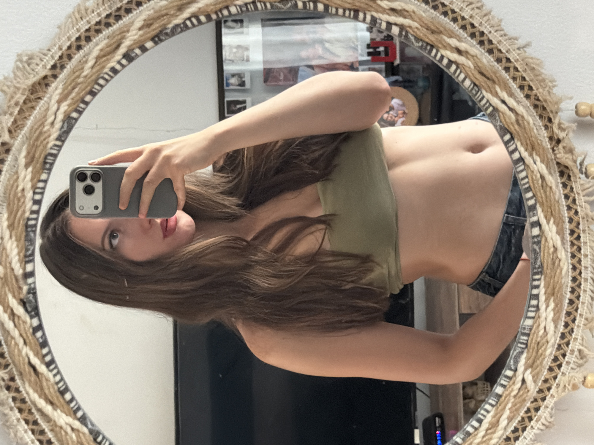 A woman is taking a mirror selfie in what appears to be an indoor setting. She is wearing a green tube top and blue jeans. Her hair is long and brown, and she has makeup on. She is holding up a smartphone to take the picture. The mirror is round with a decorative, woven frame. In the background, a television is visible, along with some framed pictures. The scene does not provide any specific indicators of a location in Sharm El Sheikh, Egypt.