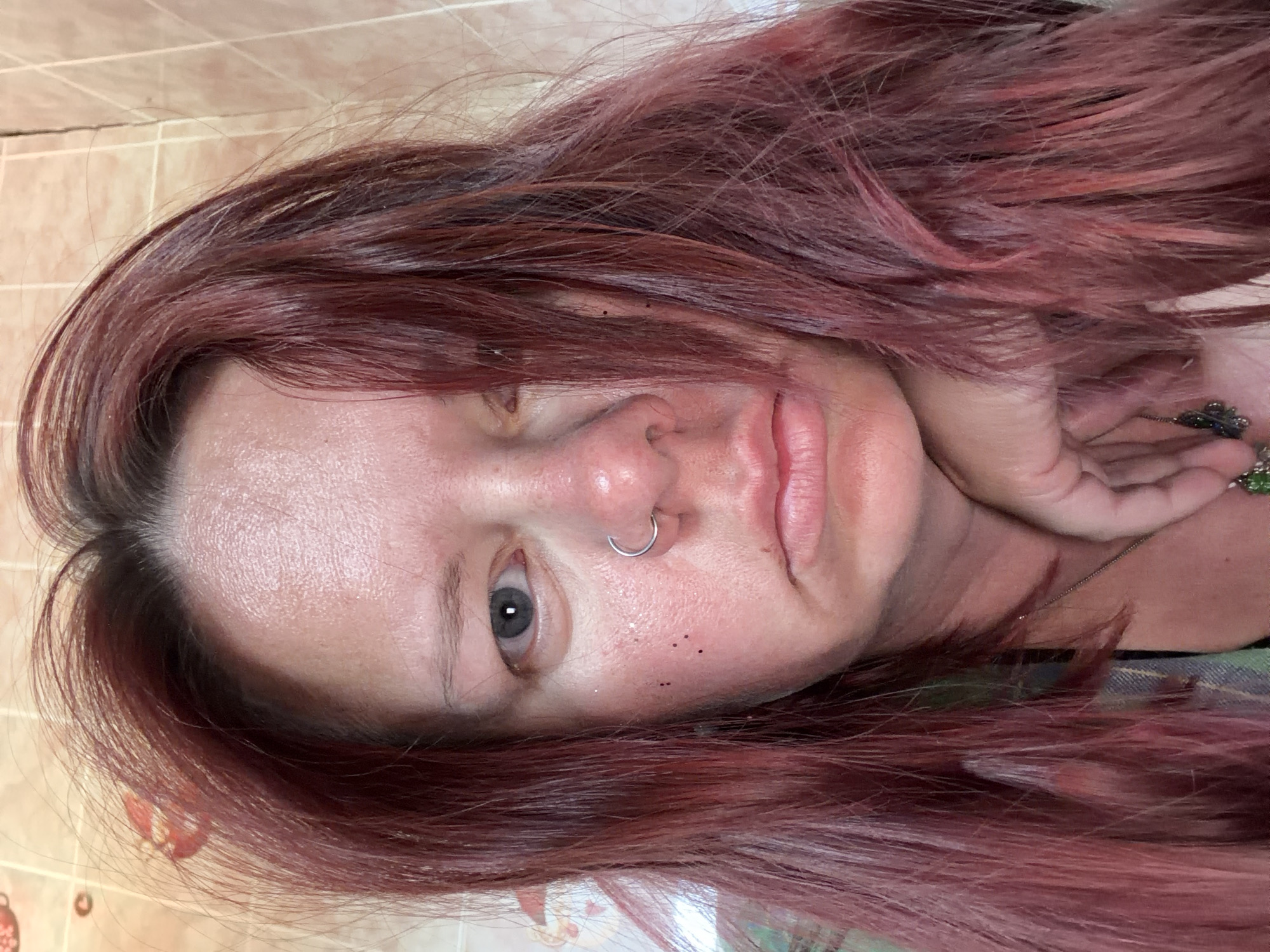 A close-up, slightly angled, selfie captures a fair-skinned individual with reddish-brown hair. The individual has a nose ring and some small dark spots or flecks on their face. One eye is visible, appearing greyish-blue. Part of a hand rests near their face, and a bit of what looks like a pendant or accessory is visible near their neck. The background appears to be a wall with light-colored, possibly tiled, surface. The photograph's location is unknown, including whether it was taken in Chaikovsky, Russia.