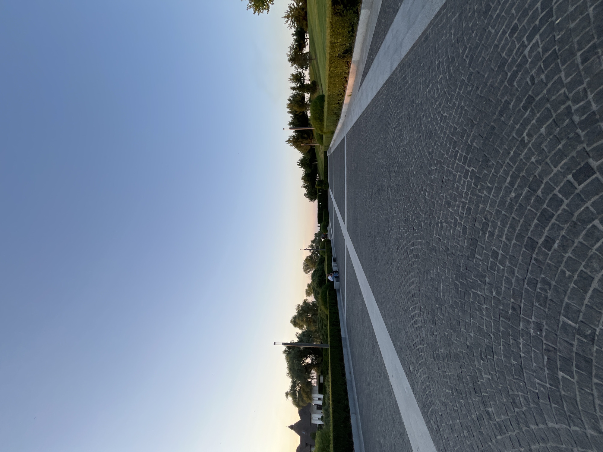 An outdoor shot captures a perspective view of a paved walkway made of dark cobblestones, with a white stripe running down the center. The walkway leads towards a serene landscape with manicured green hedges and trees. In the distance, there's a hint of a building with a distinctive roofline. Two individuals are seated on a bench along the walkway, facing away from the camera, suggesting a moment of quiet contemplation. The sky above is a clear, soft blue, indicative of early morning or late evening light. The scene is likely situated in Dnipro, Ukraine, given its characteristic urban park design and landscaping.