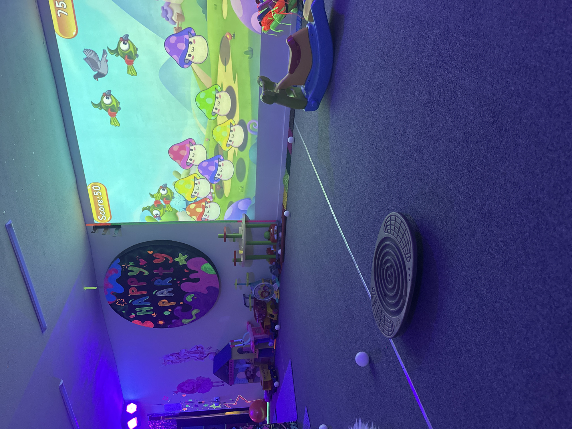 This image captures a brightly lit indoor play area, possibly a children's party venue, illuminated by vibrant purple and blue lights. A large projection screen dominates the left side of the scene, displaying a whimsical cartoon game with animated green fish and colorful mushrooms. The score "75" is visible at the top of the screen, with "Score: 50" below.
The floor is covered in a dark blue carpet. Scattered across the floor are several white balls, suggesting an interactive activity. A portion of a children's ride-on toy, a rocking horse with a purple base and a beige seat, is visible near the screen.
On the wall, a circular sign with the text "HAPPY PARTY" written in colorful, bubbly letters is prominently displayed. Below the sign, there are illustrations of figures, possibly fairies or mermaids, and star motifs, all rendered in a style that appears to glow under the dark lighting.
Towards the right of the image, a circular balancing board with a maze-like pattern is positioned on the carpet. Further back, a collection of children's toys, including what appear to be building blocks and a small playhouse, are neatly arranged. The overall atmosphere is playful and energetic, indicative of a space designed for children's entertainment. The time of day and weather are not discernible, but the artificial lighting suggests an indoor setting, possibly used during evening hours or in a space designed to be dimly lit with colored lights.