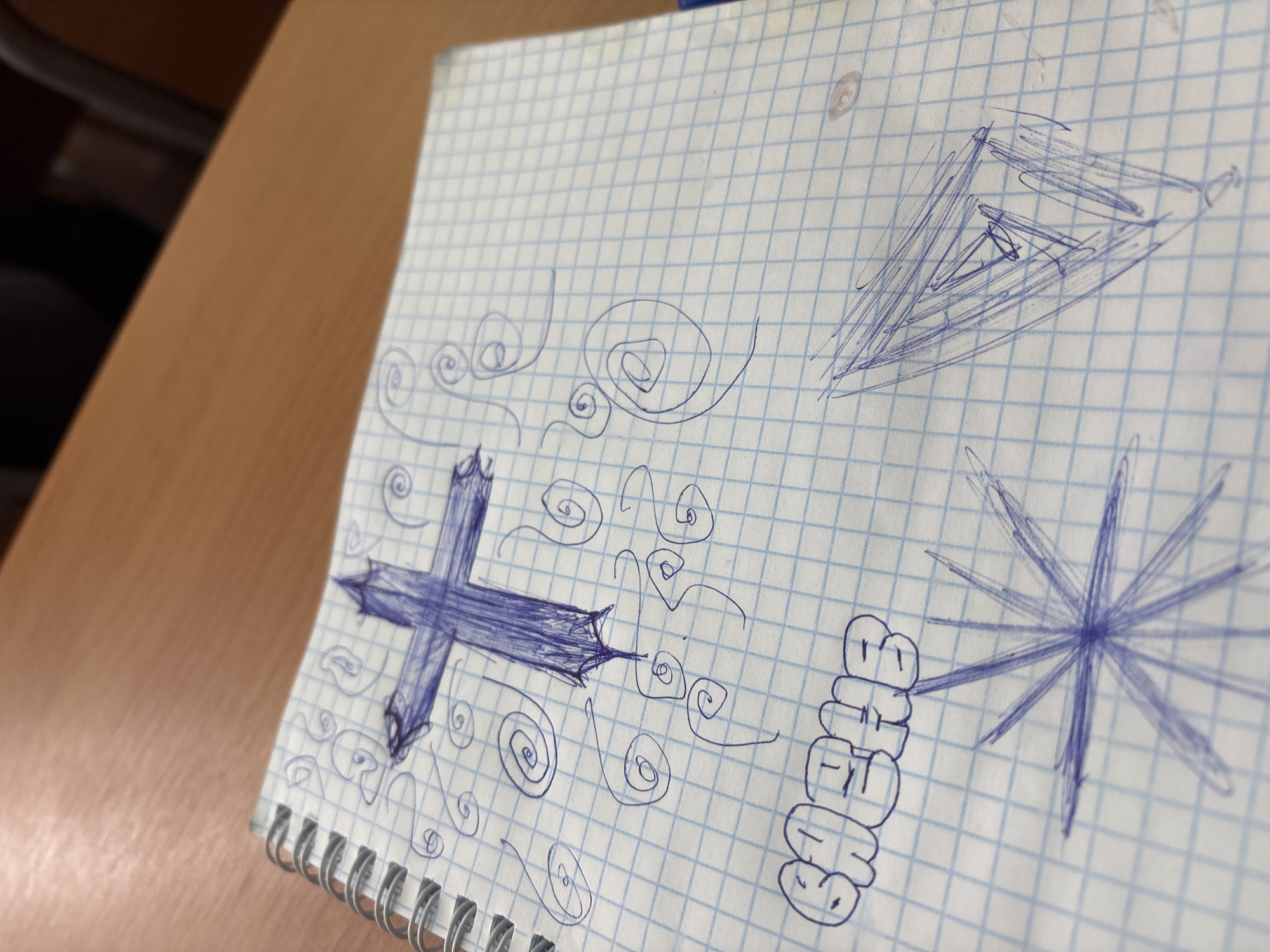 A close-up, angled shot captures a spiral-bound notebook with grid paper resting on a wooden surface, likely a desk or table. The image is taken from an overhead perspective, looking down at the notebook. The left side of the notebook and the wooden surface are closer and slightly more in focus.
The white grid paper is covered with various doodles drawn in blue pen. Prominently featured on the left side is a large, stylized cross or plus sign with sharp, pointy ends, filled in with dense blue strokes. Swirly lines and smaller spirals emanate from and surround this central doodle. Scattered across the page are more free-form spiral and wavy line patterns.
In the top right portion of the notebook, an abstract, triangular or arrow-like shape is drawn, also filled with multiple blue pen lines. Towards the bottom right, a starburst or spiky circular shape is visible, created by numerous lines radiating from a central point. Below this starburst, arranged vertically, is the word "SHELBY" written in a distinct, bubbly font.
No people are visible in the image. The activity depicted is doodling or casual drawing on notebook paper. The lighting suggests an indoor setting, but the time of day or weather cannot be determined. No specific emotions are discernible from the static image of doodles.