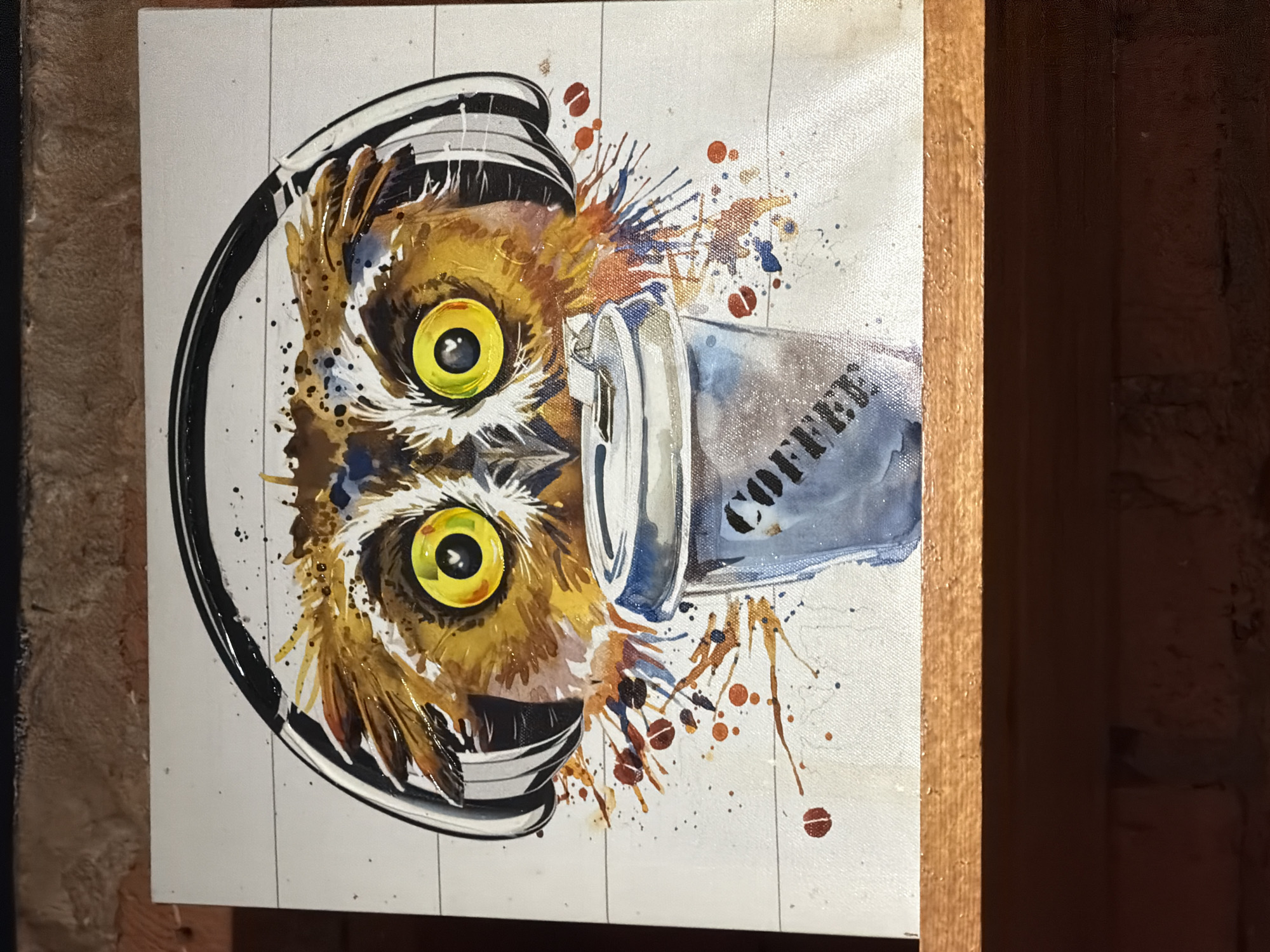 An upright, rectangular piece of artwork is displayed, resting on a light brown wooden shelf. The artwork depicts a stylized owl with prominent bright yellow eyes and black pupils, wearing black and white over-ear headphones. A silver-blue disposable coffee cup, with the word "COFFEE" written vertically in black, is positioned in front of the owl's lower face. The owl's feathers are rendered in shades of brown, orange, and black, utilizing a watercolor or paint-splatter technique, with splatters extending onto the white background. The white background of the artwork features faint horizontal lines, suggesting a plank-like texture. The wooden shelf exhibits minor surface texture. Behind and below the shelf, a dimly lit dark reddish-brown brick wall is visible, with horizontal brick courses and visible mortar lines. No identifiable features within the image indicate a specific location in Moscow, Russia.