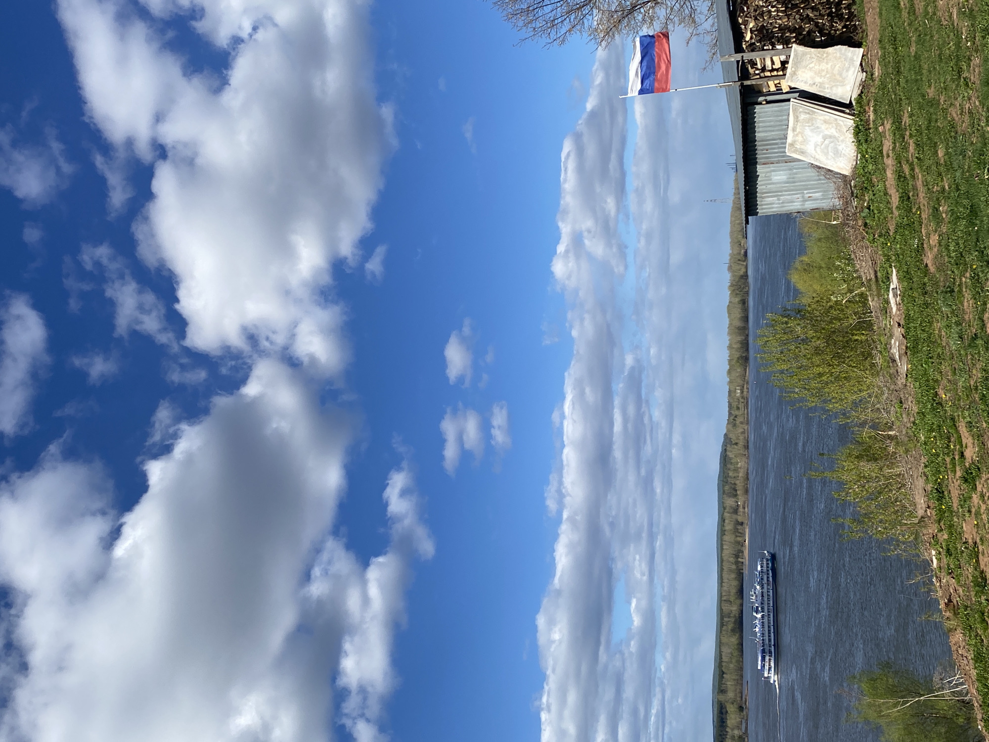 The image depicts a landscape with a clear blue sky filled with puffy white clouds on the left side of the frame. On the right side, there is a river with a passenger ship traveling along it. The riverbanks are lined with green vegetation and trees. At the top right, there is a building with a Russian flag waving from a pole. The building is surrounded by a stack of firewood and some boards. The image is likely taken in Ольховка, Russia, given the presence of the Russian flag.