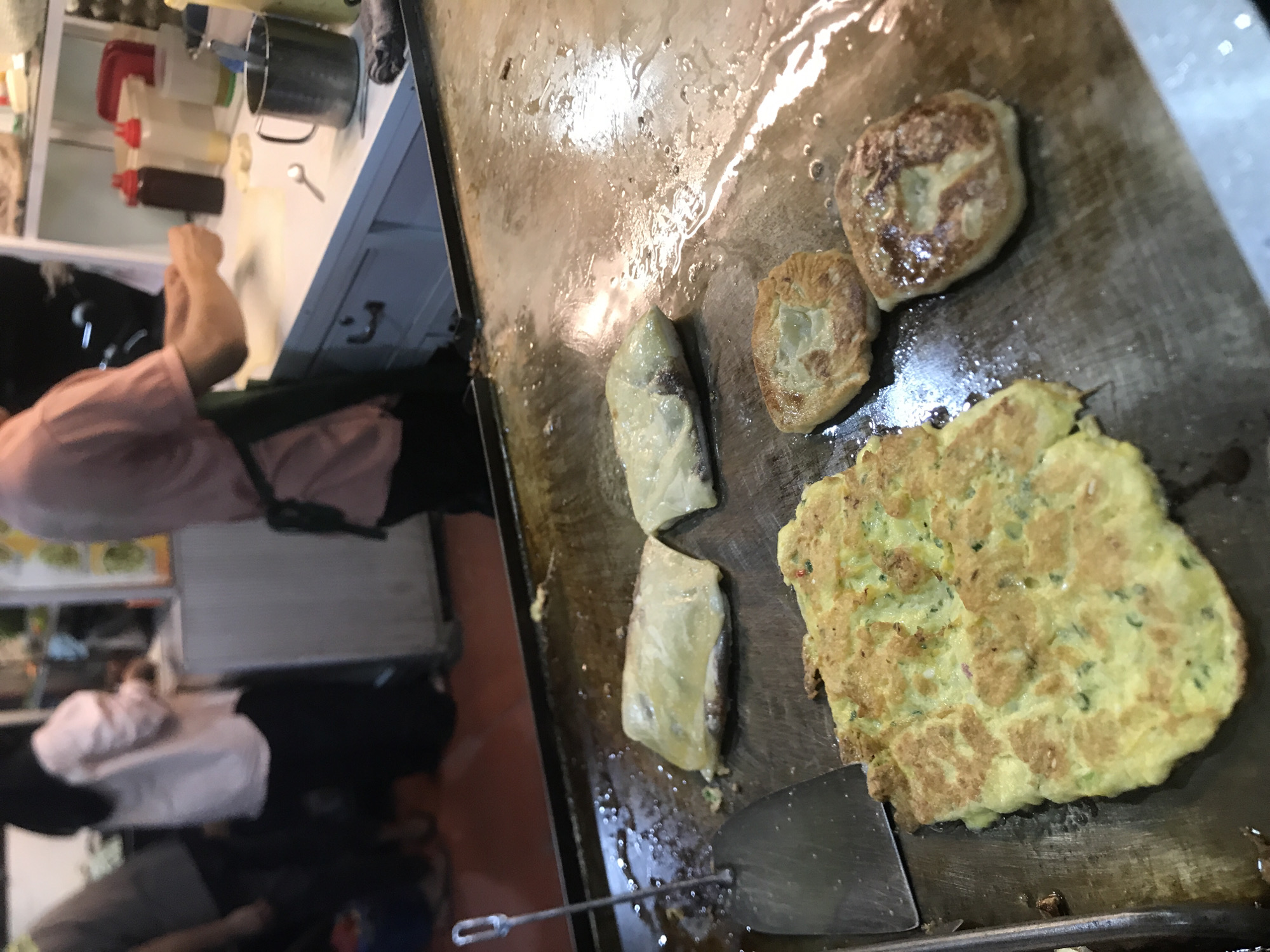The image depicts a street food stall in Lhokseumawe, Indonesia. Several food items are cooking on a flat metal griddle, including two round fried pastries, two wrapped parcels, and a large omelet. A metal spatula rests beneath the omelet. To the left of the griddle, parts of a person are visible, including a pink shirt and legs, along with portions of kitchen equipment and storage. The scene appears to be indoors under bright lighting.
