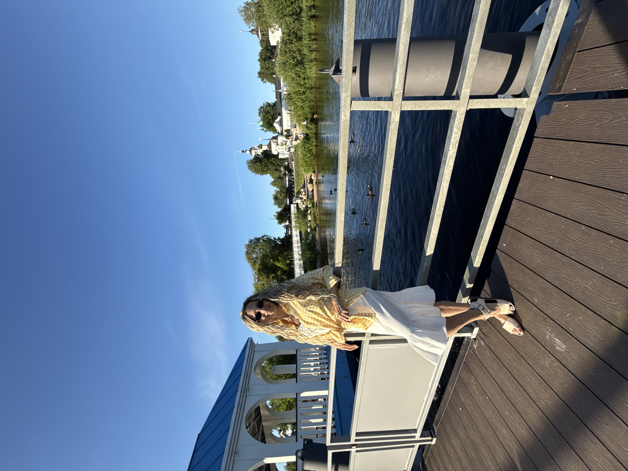 The image depicts a bright, sunny outdoor scene set on a wooden pier, described as being in Dumino, Russia. A woman with long, wavy blonde hair and round sunglasses stands in the foreground, leaning with her left arm on a grey metal railing that borders a body of dark blue water. She is smiling towards the camera. She wears a yellow and white gingham button-down shirt tied at the waist over a white tank top, paired with a white tiered midi skirt and light-colored flat sandals with ankle straps.
The pier is constructed from wide, brown composite decking planks. To the left, a white gazebo-like structure with a blue, sloped roof is partially visible. A black bucket or container rests on the pier near the gazebo.
Across the body of water, a prominent white architectural complex, characteristic of a monastery with multiple domes (some green, some dark/silver) and bell towers, is situated on the far bank. This complex is surrounded by lush green trees and bushes. Several small, indistinct figures, including what appear to be children, are visible on the distant bank near the water's edge. Approximately ten dark-colored ducks are scattered and swimming in the water between the pier and the far bank. The sky above is clear and bright blue, indicating fair weather. Strong sunlight casts distinct shadows from the railing onto the pier.