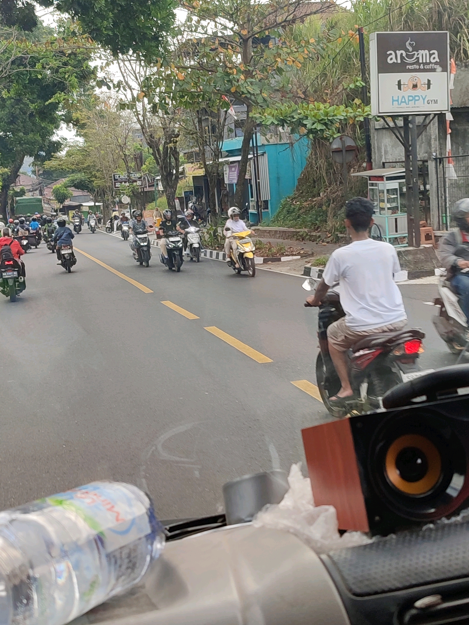 A medium shot captures a busy street scene in Wonosobo, Indonesia, focusing on a procession of motorcycles and riders. The foreground reveals a partially visible car dashboard with a water bottle and a speaker. The road is paved and marked with yellow lines, indicating traffic lanes. In the mid-ground, several motorcycles are in motion, some carrying one or two riders. Helmets are visible on many individuals, suggesting adherence to traffic safety. The riders are dressed in casual clothing, with a man in a white t-shirt and beige shorts on a dark motorcycle being a prominent figure. A woman on a yellow scooter is also present. The background showcases a street lined with buildings, trees, and a large sign advertising "aroma resto & coffee" and "HAPPY GYM". The overall environment suggests an urban setting with moderate traffic and commercial establishments. The lighting indicates it is daytime, possibly overcast given the diffused light.