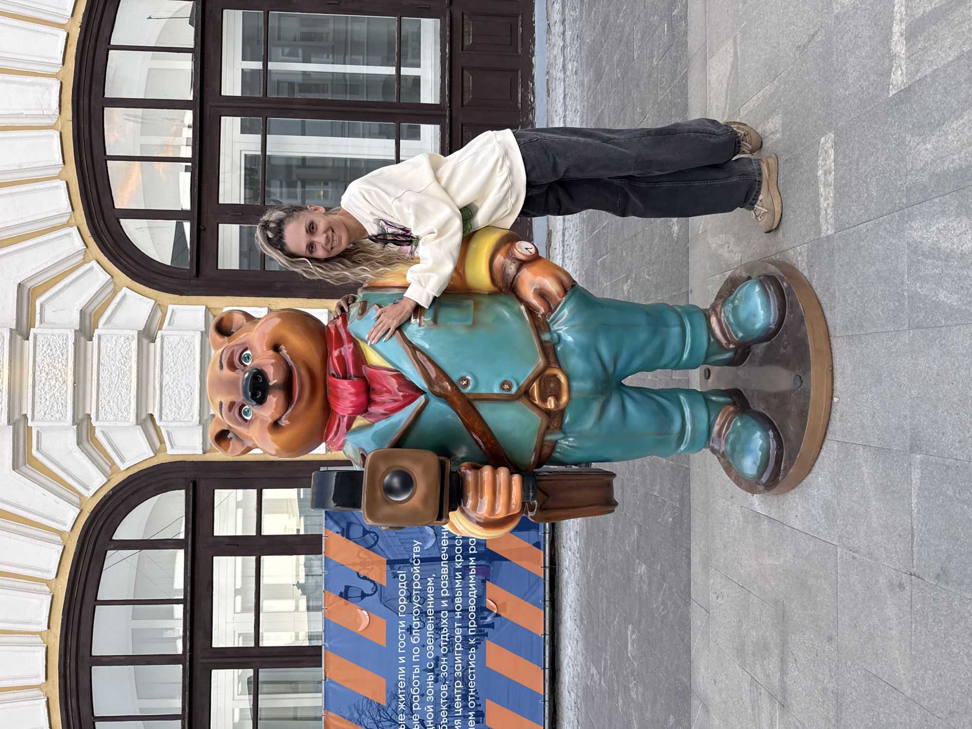 A woman with blonde hair in a ponytail is standing next to a statue of a bear. The bear is wearing a teal suit, a red scarf, and holding a vintage camera. The woman is wearing a cream-colored sweatshirt and dark jeans. She has her left hand on the bear's shoulder and is smiling at the camera. They are standing on a stone-paved sidewalk in front of a building with arched windows. A blue and orange banner with Russian text is visible in the background. The scene is likely outdoors on a cloudy day. The statue is a landmark in Rusanovo, Russia.