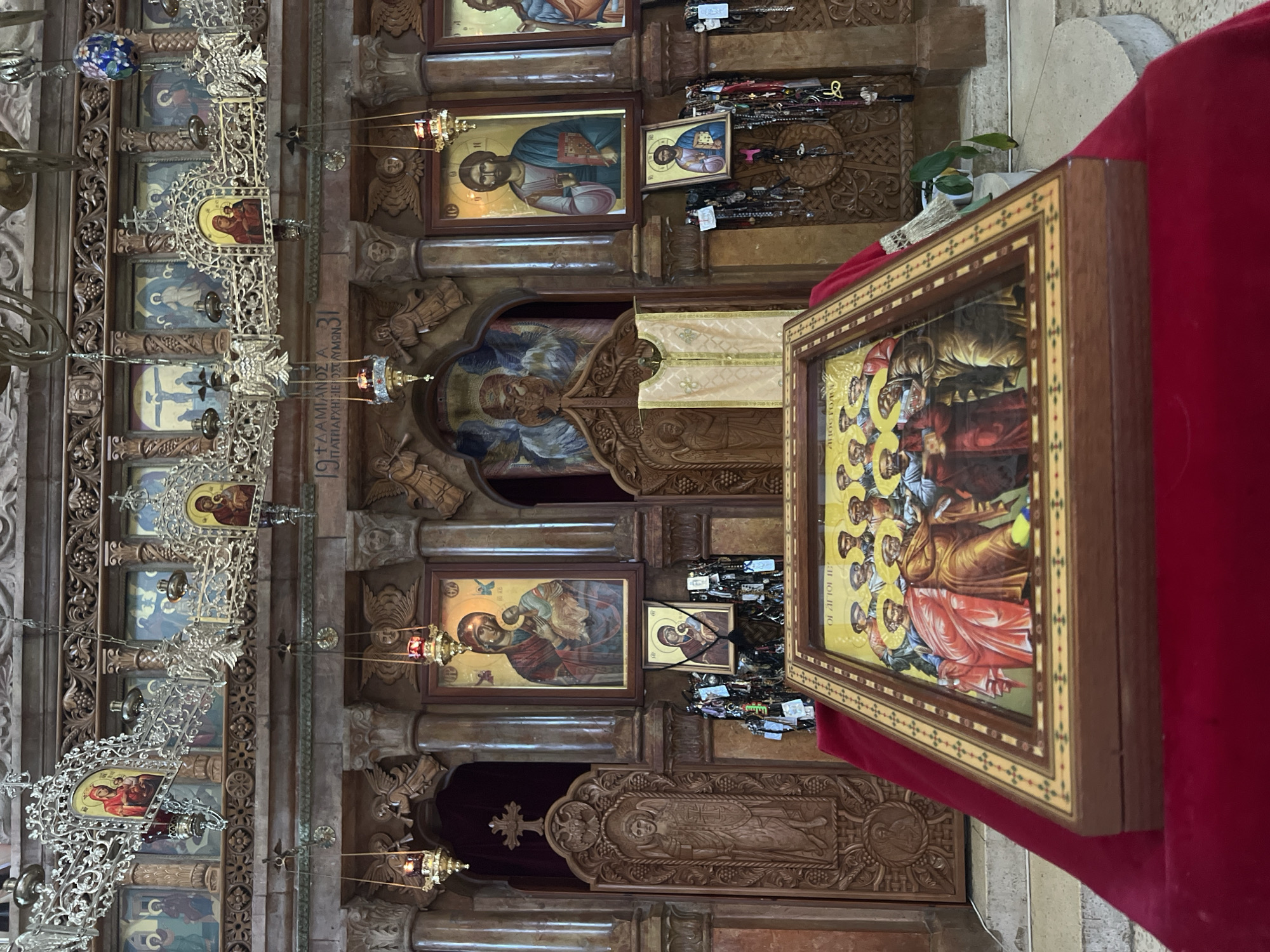 The image shows the interior of a church, likely an Orthodox Christian church, located in Emek HaYarden Regional Council, Israel. The scene is dominated by religious icons, elaborate wooden carvings, and ornate chandeliers. In the foreground, resting on a red velvet cloth, is a framed religious icon depicting a group of figures with halos. Behind this icon, a large iconostasis, a wall of icons separating the nave from the sanctuary, is visible. It features multiple icons, including images of Mary and Jesus, and Christ. The wooden structure of the iconostasis is intricately carved with religious motifs and architectural elements. Hanging from the ceiling are several metal chandeliers with oil lamps. The walls are adorned with a series of smaller icons and decorative carvings. The overall atmosphere is one of solemnity and religious devotion.