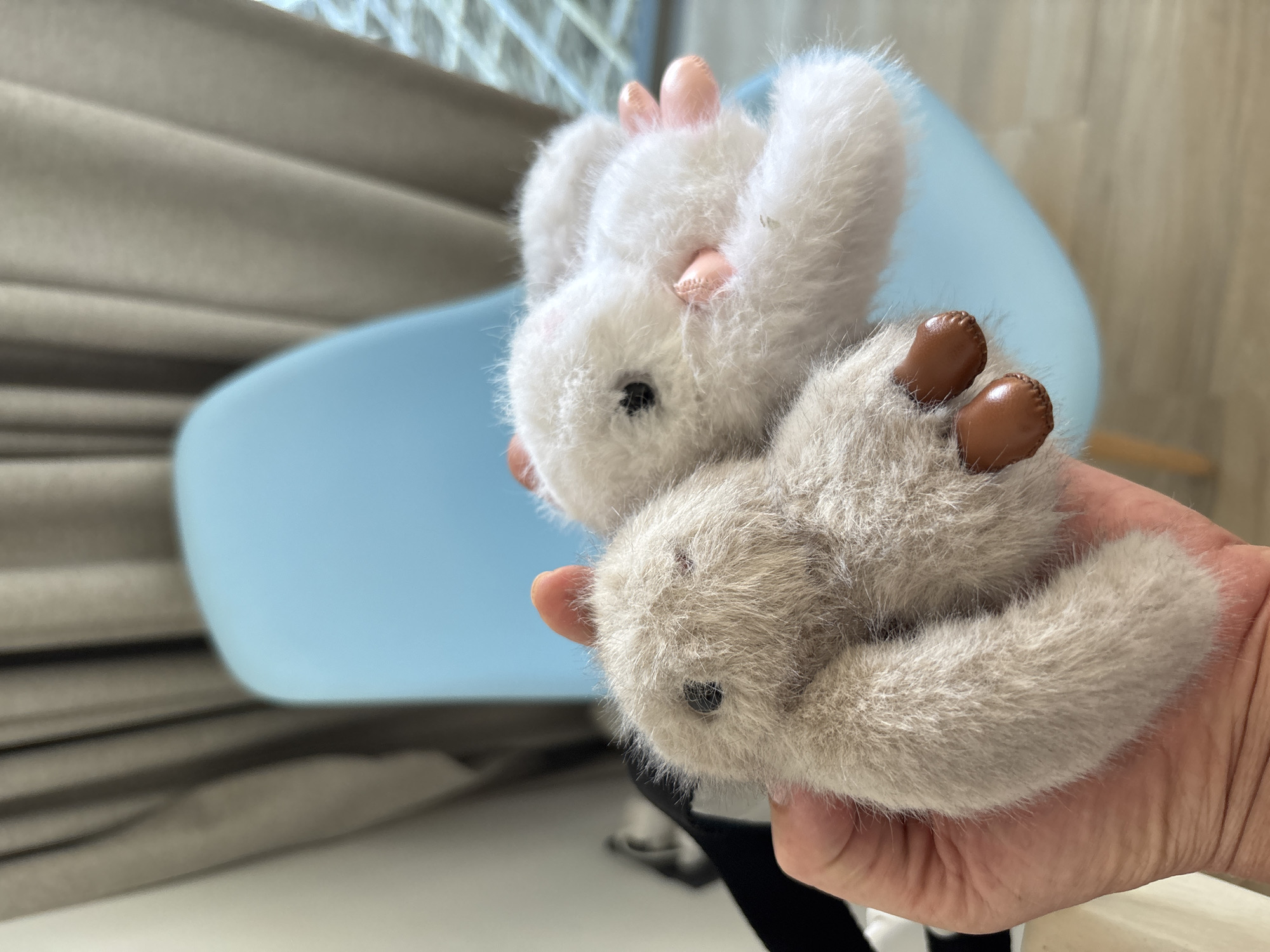 A hand holds two small, fuzzy, toy animals. The animal on top is white with pink ears and feet. The animal on the bottom is grey with brown feet. The background is blurry, but a light blue chair and a window with blinds are visible. The location is Baan Chaweng Noi, Thailand.
