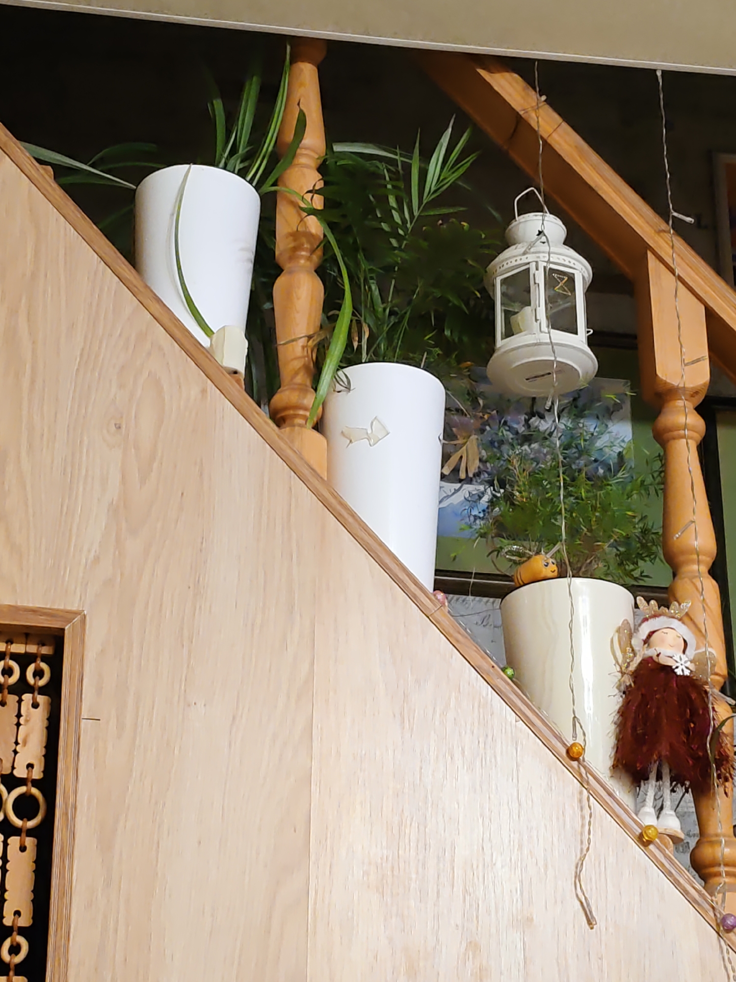 The image shows a close-up view of a wooden staircase, likely in a residential setting in Rostov-on-Don, Russia. The staircase is decorated with several potted plants in white and cream-colored pots. Hanging from the wooden banister is a white lantern and a string of small, colorful lights. A small, doll-like figure with wings and a feathered dress is also visible on the staircase. The overall impression is one of a cozy, decorated interior.