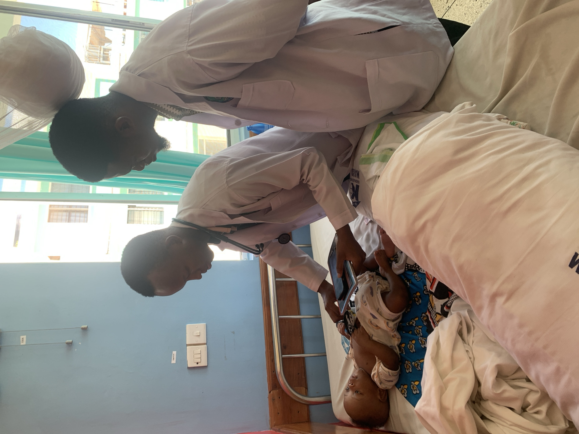 Two doctors are examining a baby in a hospital room. The doctors are both wearing white coats and stethoscopes. One doctor is holding a phone and recording the examination. The baby is lying on a bed covered with white sheets and a blue patterned cloth. The room has blue walls, and there is a window in the background. The floor is red. The location is Dar es-Salaam, Tanzania.