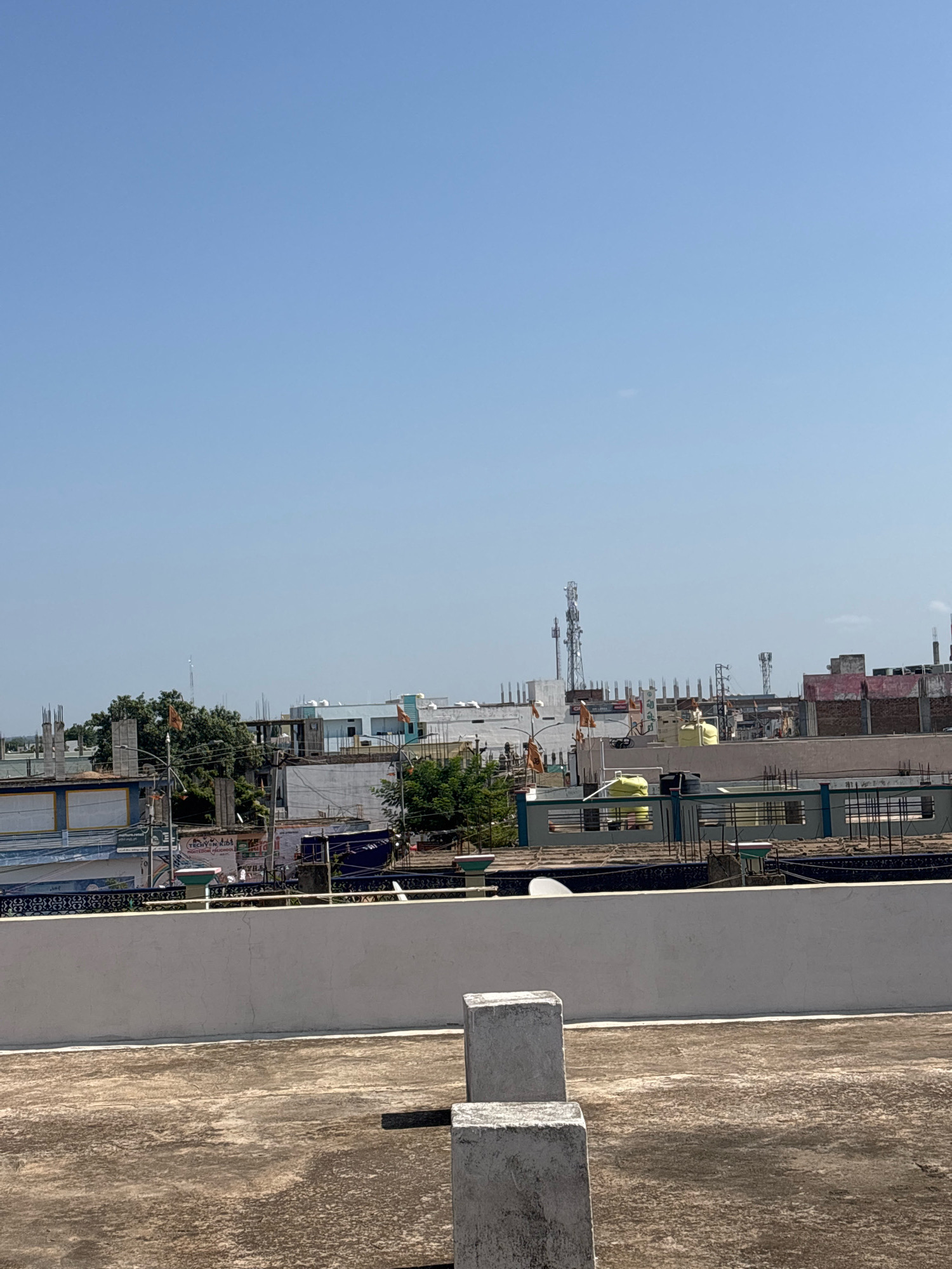 The image shows a rooftop in Aija, India, with a clear blue sky. In the foreground, a concrete slab is visible. The mid-ground features a low, white-washed parapet wall. Beyond the wall, a cityscape unfolds with various buildings, some under construction with exposed rebar. Several telecommunication towers are prominent in the background, along with water tanks on top of some buildings. Trees are scattered throughout the urban landscape. There are no people or animals visible in the image.