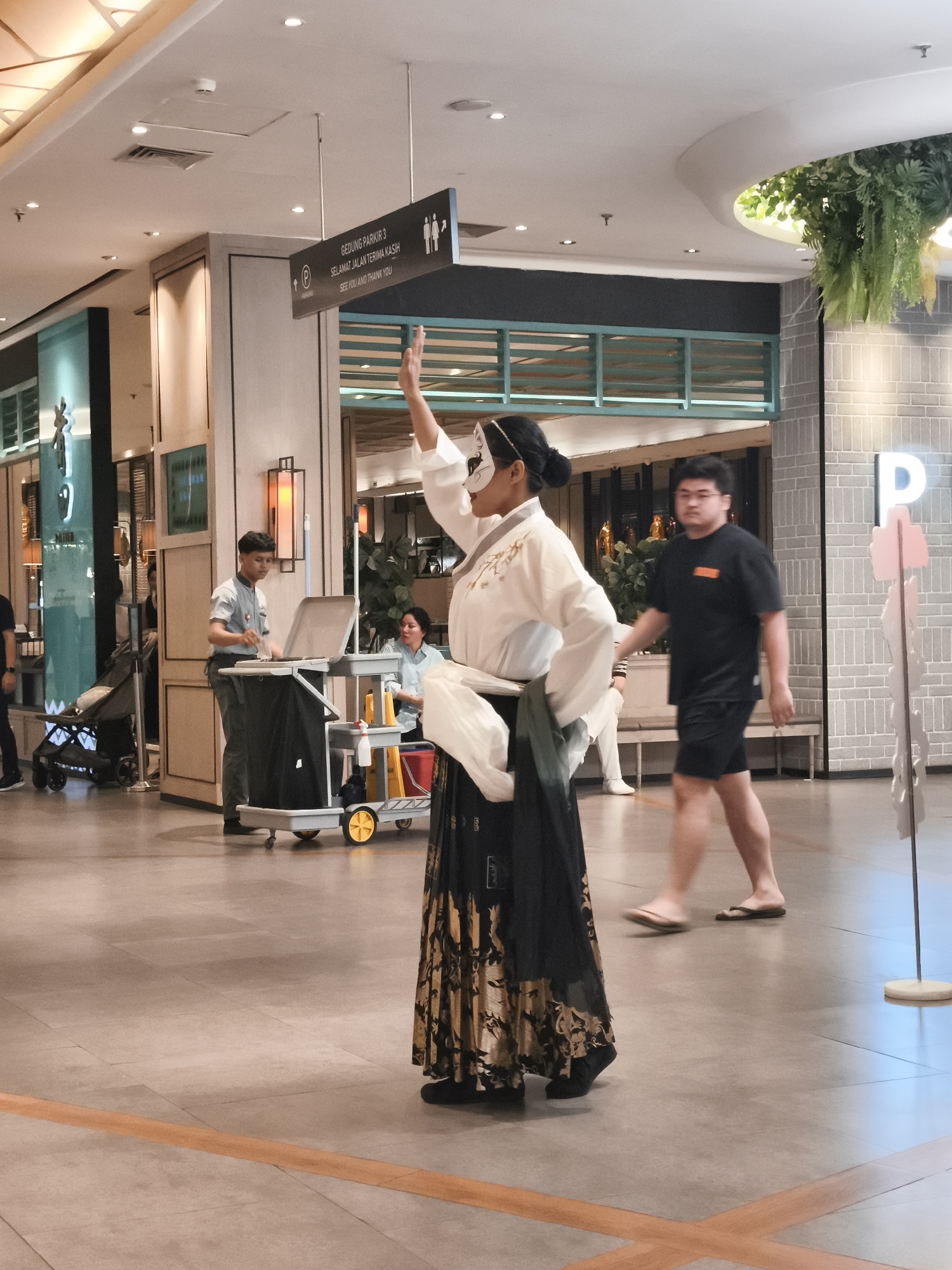 The image depicts an indoor environment, likely a shopping mall or a similar public space in North Jakarta, Indonesia. In the foreground, a person wearing a traditional costume and a white mask is performing or posing, with their right arm raised. The costume consists of a white top with embroidery and a long, flowing skirt with a gold and black pattern. Behind this performer, a young man in a black t-shirt and shorts walks past. To the left, a cleaner pushes a trolley. In the background, there are various architectural elements, including signs indicating "GEDUNG PARKIR 3" and "SEE YOU AND THANK YOU," as well as decorative plants and lighting fixtures. The floor is tiled with a large orange line marking a pathway.
