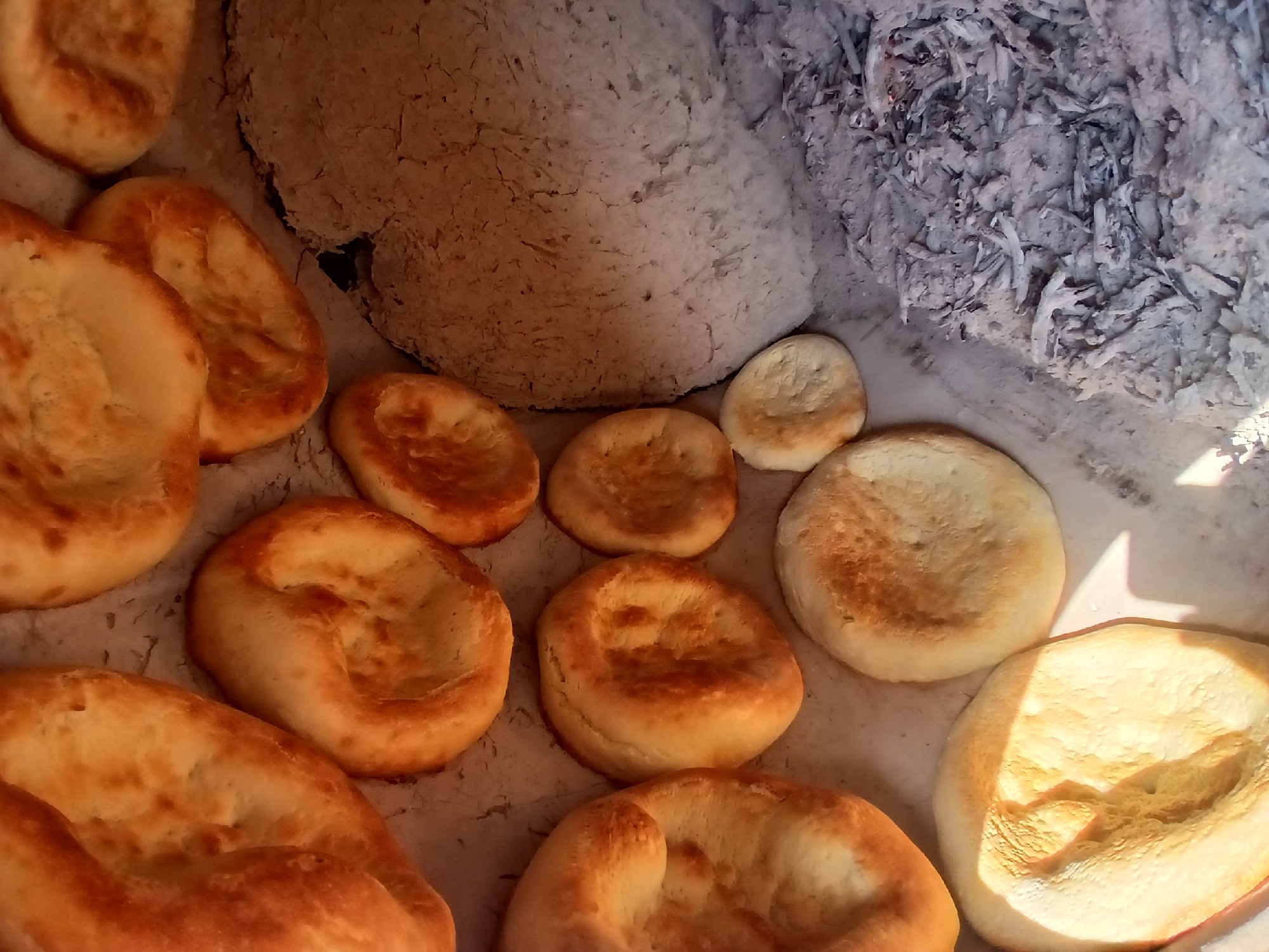 In the city of Yangiarab, Uzbekistan, an overhead shot captures an arrangement of freshly baked flatbreads placed on a traditional clay oven. Several round, golden-brown flatbreads, some with slightly darker, blistered edges, are positioned on a light-colored surface. To the right, a portion of the oven's interior is visible, characterized by its rough, grey, clay texture. At the bottom right of the image, a pile of grey ash and embers suggests the oven is either recently used or still hot. The lighting indicates an outdoor setting, likely during the day, with shadows cast on some of the bread. There are no people or animals present in the frame.