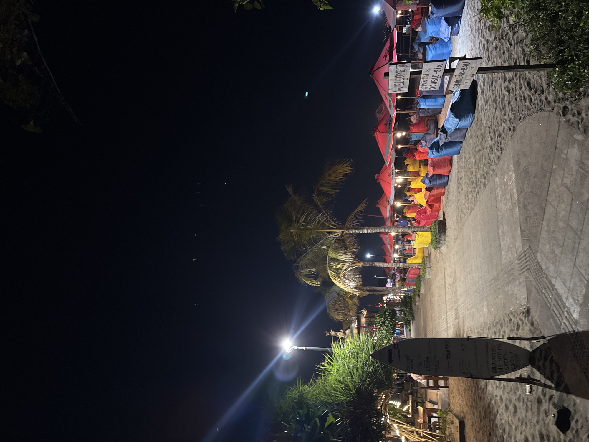 The image depicts a nighttime scene on a beach in Kuta, Indonesia, likely Seminyak Beach, given the signage. The foreground is dominated by sand with a paved walkway to the right. A surfboard leaning on a stand serves as a menu board, advertising "LEARN SURF," "COLD DRINKS," and listing various beverages. Further along the walkway, a series of brightly colored beanbag chairs are arranged in front of red beach umbrellas. Several people are seated on these beanbag chairs, interacting with each other or enjoying the ambiance. In the background, palm trees and other vegetation are visible, along with some distant lights, suggesting the presence of buildings or structures. A tall lamppost illuminates a portion of the walkway. A signpost reads "WELCOME TO PANTAI DOUBLE-SIX SEMINYAK-BALI," confirming the location. The overall atmosphere is relaxed and festive, with the warm glow of lights contrasting with the dark night sky.