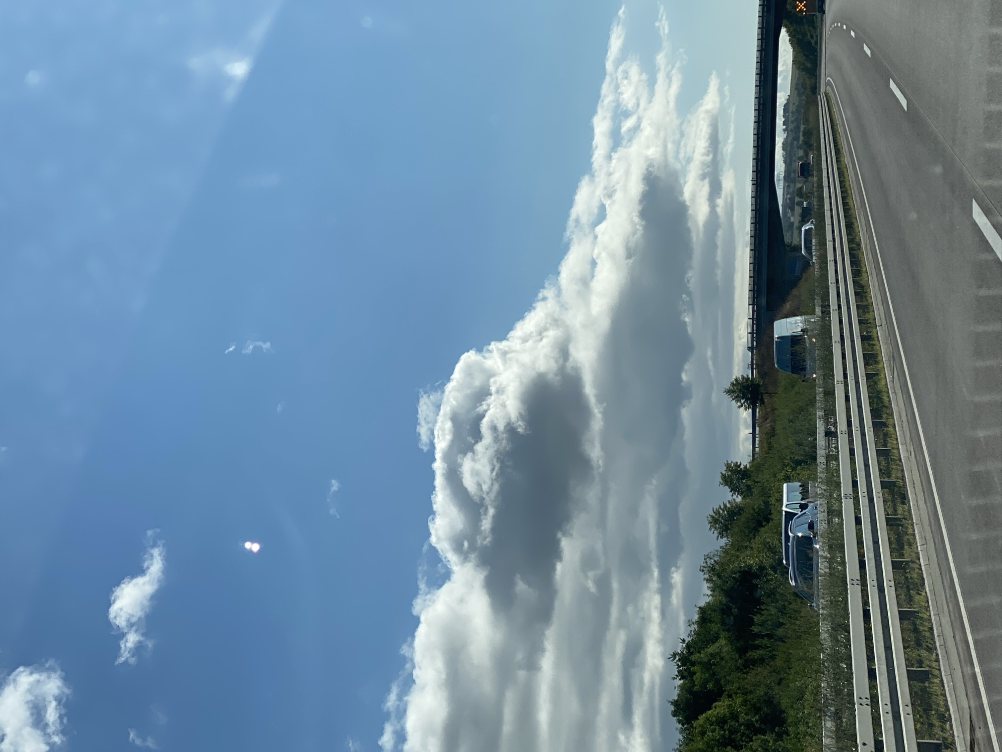 The image captures a highway scene in the vicinity of Heringen/Helme, Germany. The perspective is from a vehicle traveling on the road, looking forward. The sky is partly cloudy with large, white cumulus clouds dominating the upper portion of the frame. Two distinct bright spots are visible in the sky, possibly lens flares or reflections. A highway overpass stretches across the scene in the middle ground, with vehicles on it. The foreground shows the highway itself with lane markings and a guardrail. To the left of the highway, dense green vegetation and trees line the embankment. Several vehicles are visible on the highway, including vans and a car, some of which are stopped or moving slowly. The overall environment suggests a typical daytime scene on a German autobahn.