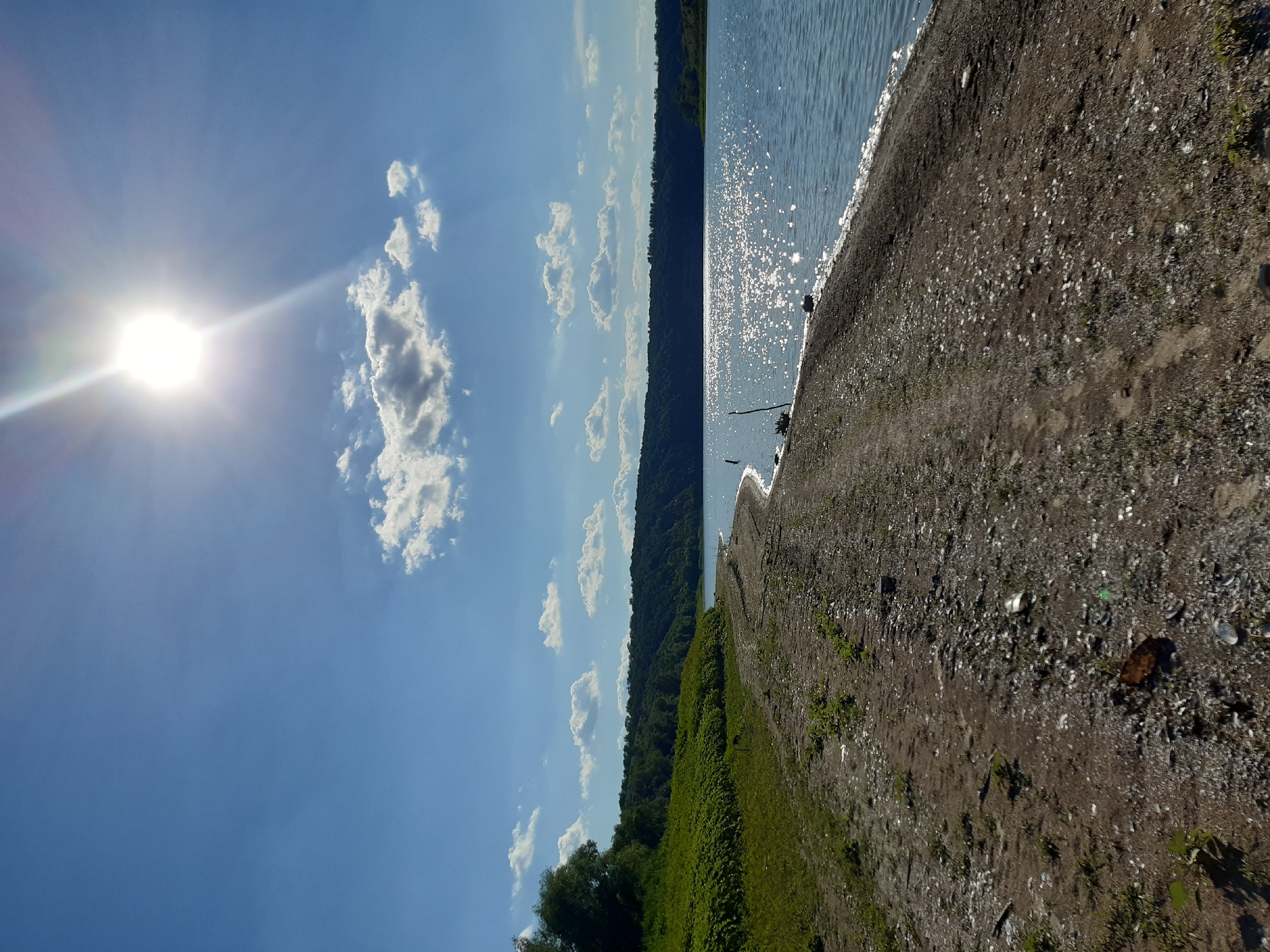 The image depicts a wide body of water, likely a river or lake, under a bright sunny sky. A dirt path runs along the shoreline, strewn with gravel and small debris. Lush green vegetation covers the sloping bank to the left, extending towards a dense line of trees and hills in the distance. The sun, positioned high in the sky, casts a strong glare on the water, creating a sparkling effect. A few scattered clouds dot the bright blue sky. The scene appears to be a natural landscape with no human presence immediately visible. The description implies this scene is located in Sokil, Ukraine, a detail that contextualizes the natural environment as being within that region.