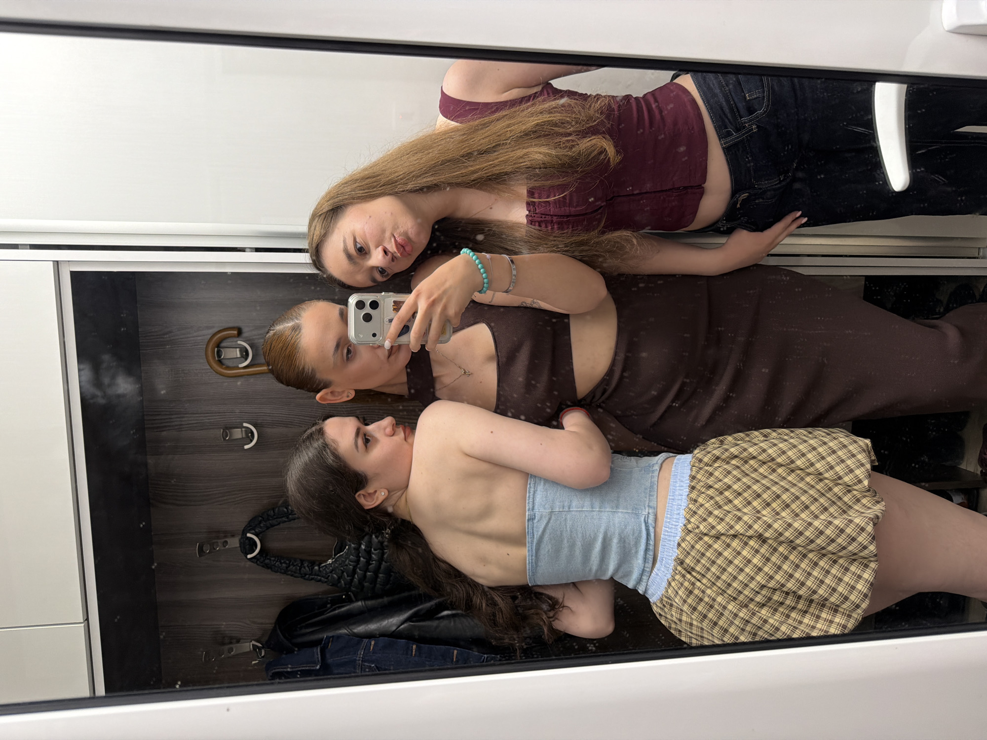 Three women are positioned in front of a mirror, presumably taking a selfie. The topmost woman has long brown hair and is wearing a maroon top and dark jeans. The middle woman is holding a smartphone, taking the photo. She has her hair in a bun and is wearing a brown dress. The bottom woman has long dark hair and is wearing a light blue top and a checkered skirt. Behind the women is a wardrobe with clothes and accessories, including a bag and some hooks. The precise location cannot be identified from the image.