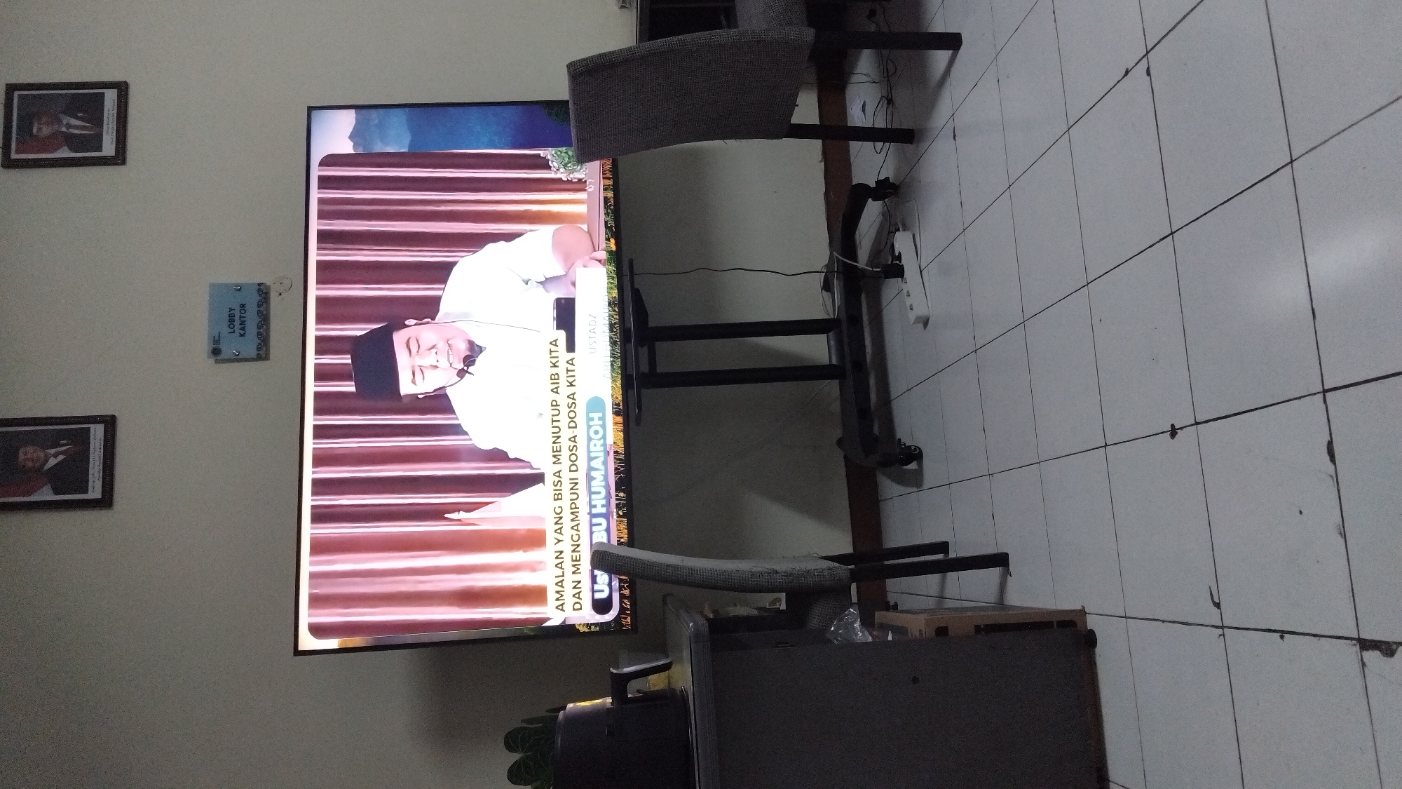 The image depicts a television screen displaying a man wearing a white shirt and a black cap, speaking into a microphone. The screen also shows text in Indonesian: "AMALAN YANG BISA MENUTUP AIB KITA DAN MENGAMPUNI DOSA-DOSA KITA" and "Ustadz BU HUMAIROH". This suggests the man is a religious speaker or cleric. The television is mounted on a stand with wheels. In the background, there are two framed portraits on the wall, a sign that reads "LOBBY KANTOR", and a chair. A desk with a plant is visible to the left of the television. The floor is tiled. There are no clear indicators placing the scene in Pekalongan, Indonesia, but the Indonesian text suggests the region.