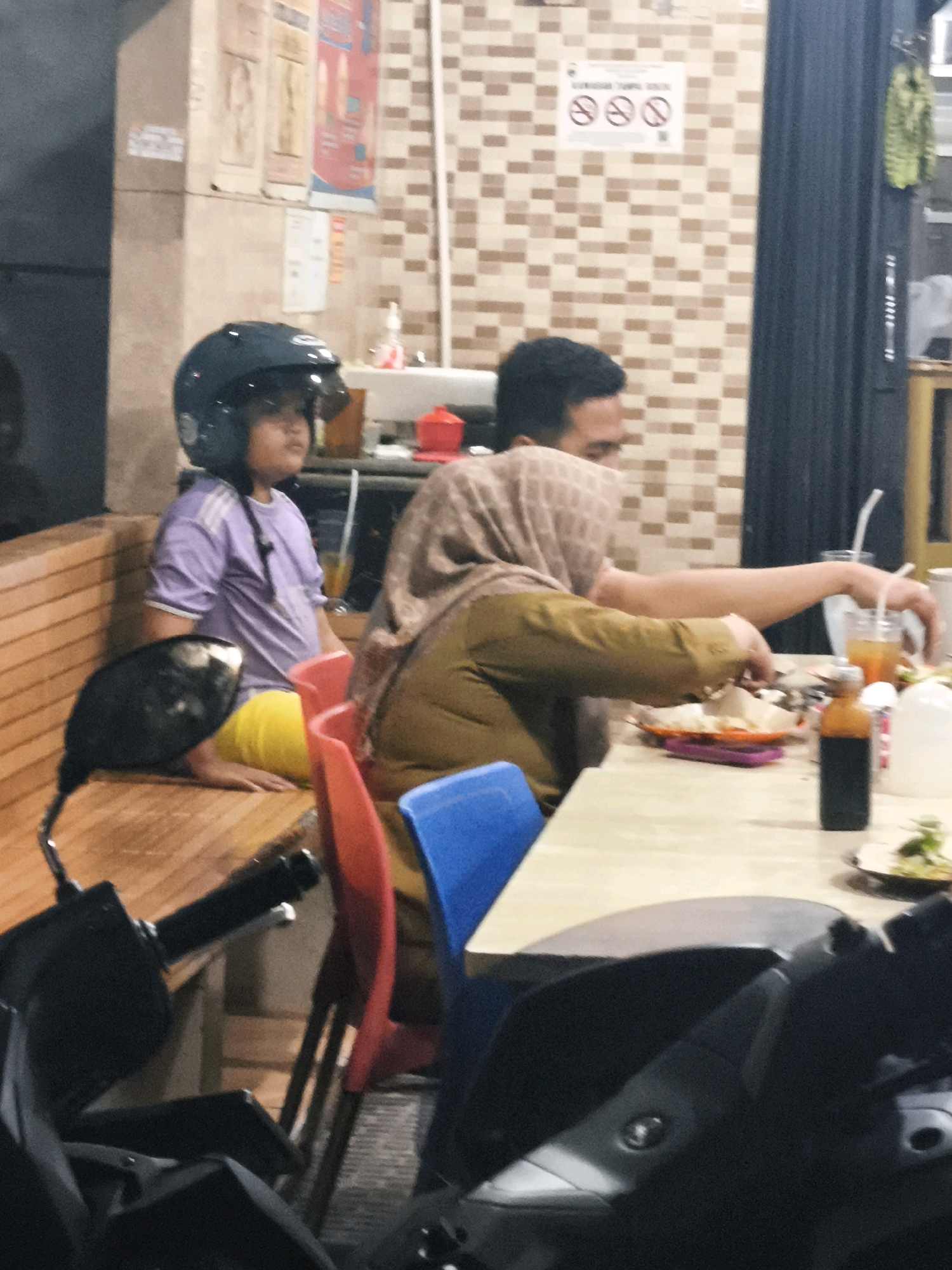 The image depicts an indoor scene, likely a food stall or casual eatery, with patrons seated at tables. A young person wearing a black helmet and a purple shirt sits on a wooden bench. Beside them, an adult wearing a headscarf and an olive-green garment is seated at a table, reaching for food. The table is set with plates of food, drinks in plastic cups with straws, and a small bottle of sauce. In the foreground, parts of a motorcycle are visible, suggesting a mode of transportation. The background features tiled walls and a counter area. The overall ambiance suggests a lively, informal dining setting in Palembang, Indonesia.