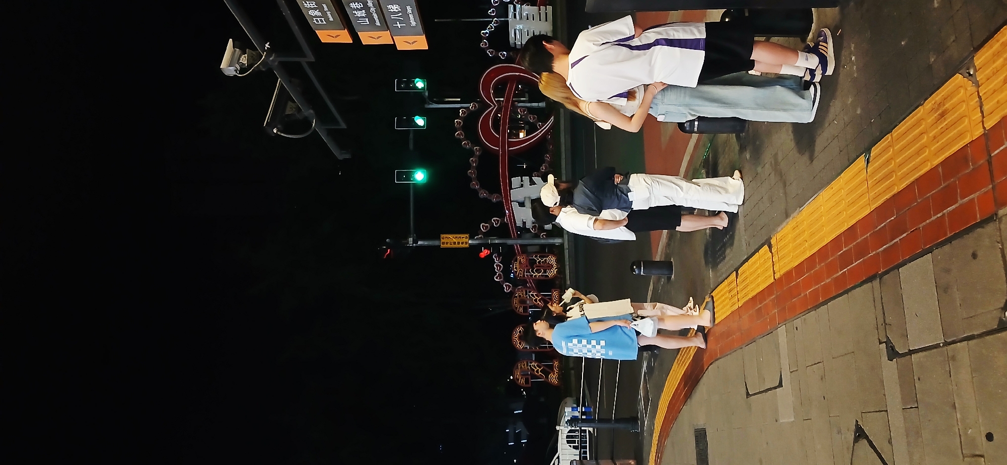 The photograph captures a night scene in what appears to be a public space in the Yuzhong District of Chongqing, China. The lighting is dim, with streetlights illuminating the scene.
In the foreground, a sidewalk marked with tactile paving stretches along the frame's right edge. Three sets of couples are present on the sidewalk. The first couple is facing away from the camera, both with their arms around each other. The man is wearing a blue patterned short-sleeve shirt and white shorts. The second couple are in the same position as the first, the man is wearing a dark-colored t-shirt with the woman wearing white. The third couple is also embracing each other, the man is wearing a black short-sleeve shirt with a white striped collar, and the woman is wearing jeans.
Beyond the sidewalk, the scene opens up to a street featuring a traffic light showing green. A decorative display resembling a Ferris wheel and heart shapes provides a backdrop, adding a festive or romantic element to the setting. Street signs can be seen above the traffic lights, with the names of nearby streets written on them in Chinese. The names of the streets written on the signs are "Raising Street", "Mountain City Alley", and "Eighteen Steps".
The overall environment is urban, and the photograph captures a moment of people enjoying the evening in a public space.