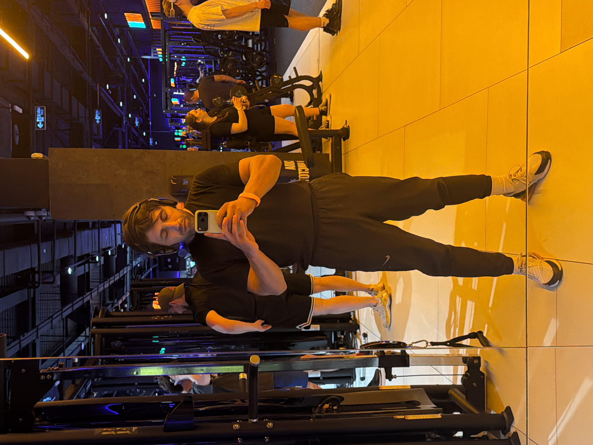 The image depicts the interior of a gym in Krasnoyarsk, Russia. A man wearing a black shirt, black pants, and sneakers is taking a selfie with his smartphone. He also wears headphones and a pink wristband. The scene includes other gym-goers using various machines. Gym equipment such as treadmills and weight benches is visible. The floor appears to be reflective. The lighting in the gym is a combination of bright white and blue.