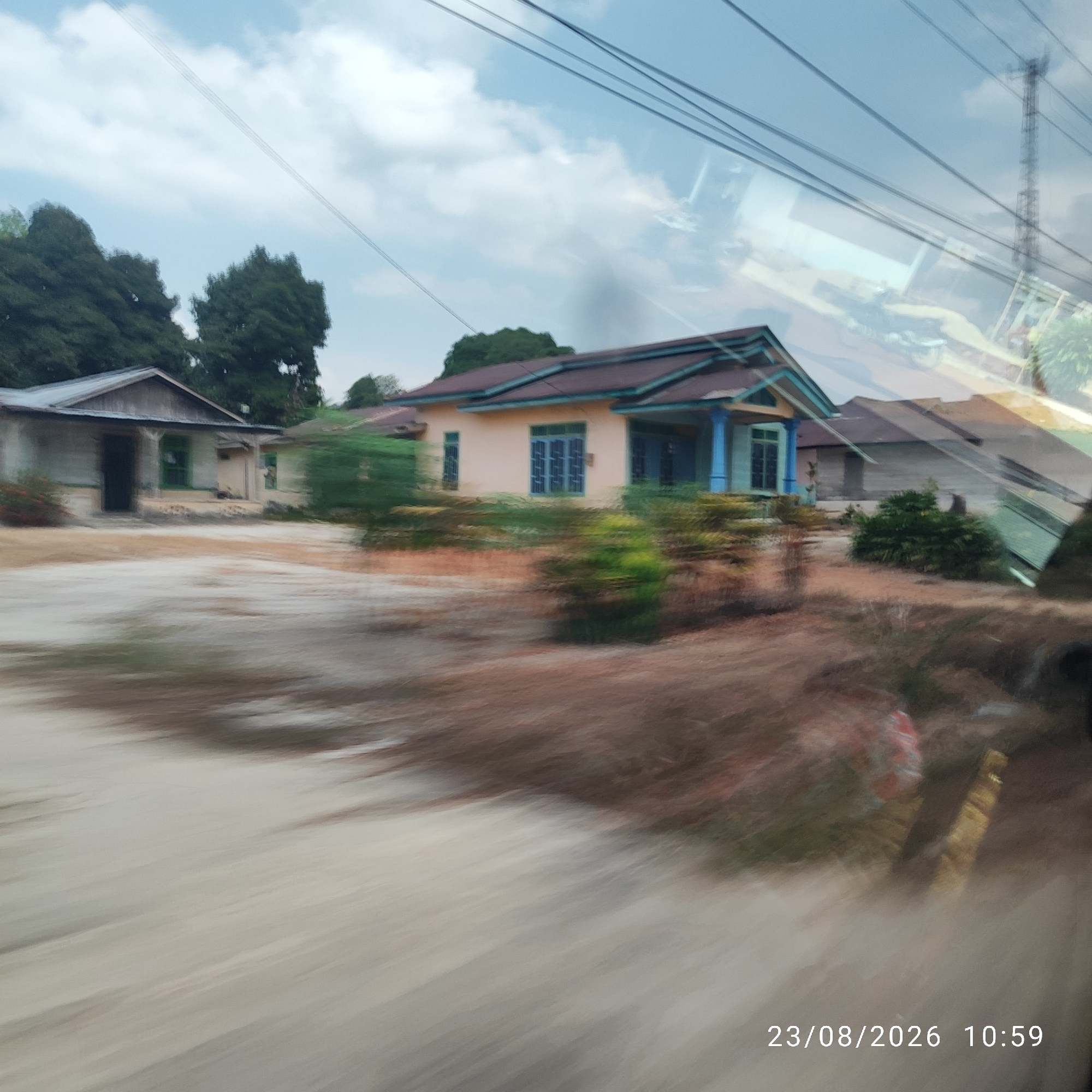 The image displays a blurry, long-exposure photograph of a residential area, likely taken from a moving vehicle. The scene captures a street with several houses and vegetation. The main subject is a light-colored house with a brown roof and blue window frames, featuring a porch supported by blue pillars. To the left of this house is another smaller, older-looking structure with a grey roof. The ground is a mix of sandy dirt and dry grass. Overhead, power lines stretch across the frame, with a cell tower visible in the upper right. The sky is partly cloudy and blue. A timestamp in the lower right corner indicates "23/08/2026 10:59". The blurriness suggests motion, implying the photograph was taken while traveling. This scene is located in Kacang Botor, Indonesia.