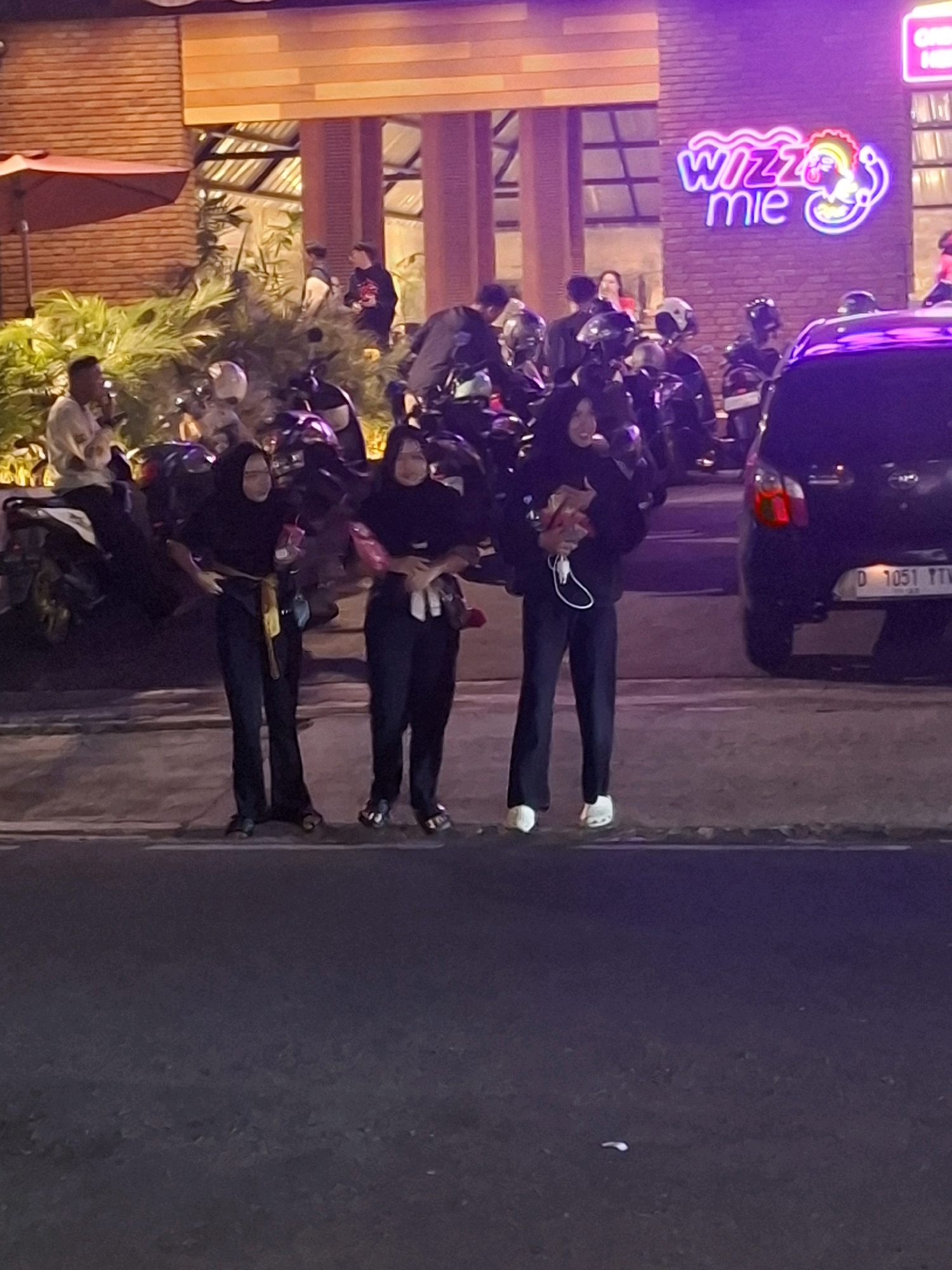 Three young women are standing on a sidewalk next to a street at night. They are all wearing dark hijabs and long pants. The woman on the left is holding a paper bag and a red drink. The woman in the middle is holding a red drink and a paper bag. The woman on the right is also holding a red drink and a paper bag. Behind them, there are several motorcycles parked and a black car with a license plate that reads "D 1051 YTV". A neon sign that says "Wizzz mie" is visible on the building behind the women. The building appears to be a restaurant or cafe. The overall lighting suggests it is nighttime, possibly in an urban environment within Cianjur, Indonesia.