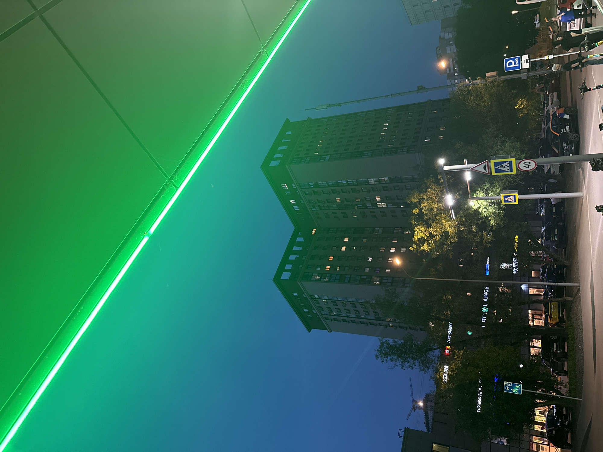 The image shows a nighttime cityscape in Moscow, Russia, with a tall, multi-story building illuminated by interior lights in the center. Streetlights cast a glow on the surrounding trees and street furniture, including road signs and parked vehicles. A large, green, artificially lit structure dominates the left side of the image, potentially part of a building or signage. A construction crane can be seen in the distance, suggesting ongoing development in the area. The sky is a gradient of dark blue, indicating early evening or late night. There are no visible people in the image.