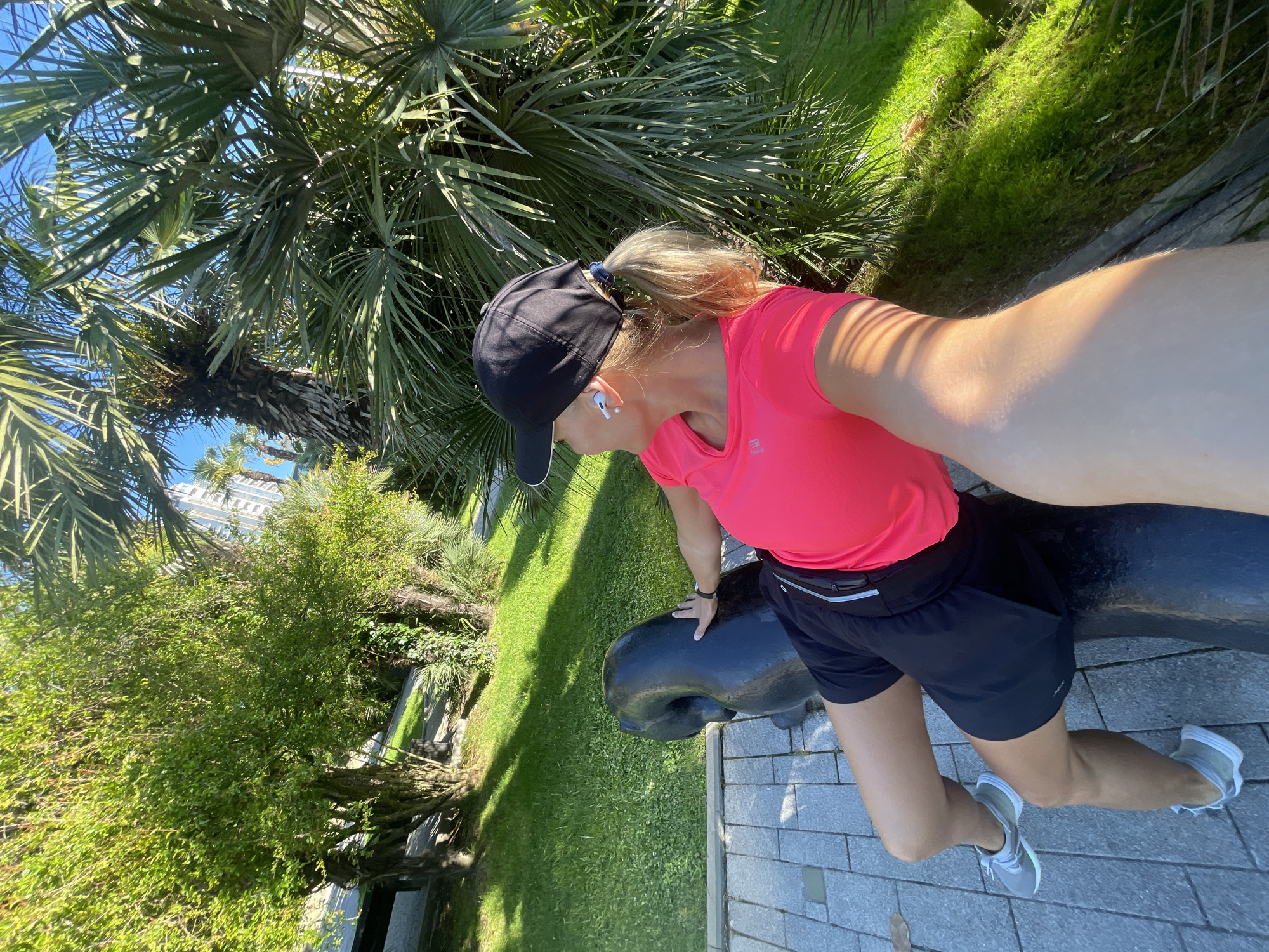 A female individual is captured in a high-angle, tilted outdoor selfie, suggesting a dynamic perspective. She wears athletic attire, including a black baseball cap, a bright coral-pink short-sleeved top with the brand "KALENJI" and its logo visible, black athletic shorts, and grey and white athletic shoes. Her blonde hair is tied in a ponytail, peeking out from under her cap, and she has white wireless earbuds in her ears. She is leaning with her left hand resting on a large, dark, rounded object that resembles a decorative cannon or sculpture, positioned on a grey paved walkway.
The setting is a lush, sun-drenched park or garden, consistent with the subtropical climate of Sochi, Russia. Abundant green vegetation surrounds her, with numerous tall palm trees dominating the background and overhead, their fronds splaying against a clear blue sky. Bright green grass borders the paved path. In the far distance, partially visible through the foliage, is a tall, light-colored building, possibly a hotel or apartment complex. The bright sunlight casts distinct shadows, indicating a clear, sunny day, likely morning or early afternoon. The overall impression is one of an active moment in a pleasant, verdant outdoor environment.