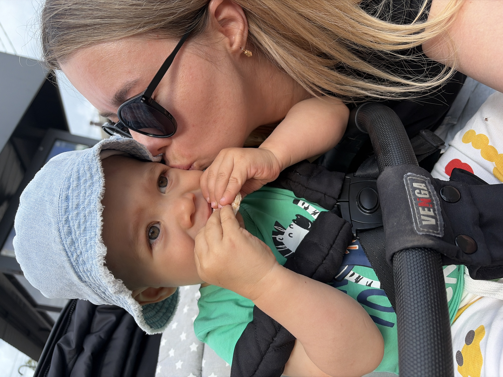 In Chaikovsky, Russia, a mother is kissing her baby on the cheek. The baby, wearing a light blue denim bucket hat and a green t-shirt with a zebra print, is sitting in a stroller and eating a cracker. The mother, with blonde hair and black sunglasses, is leaning in to kiss the baby. The stroller has black straps and a handle with the brand name "VENGA" visible. The background suggests an outdoor setting, possibly near a building with a dark roof.
