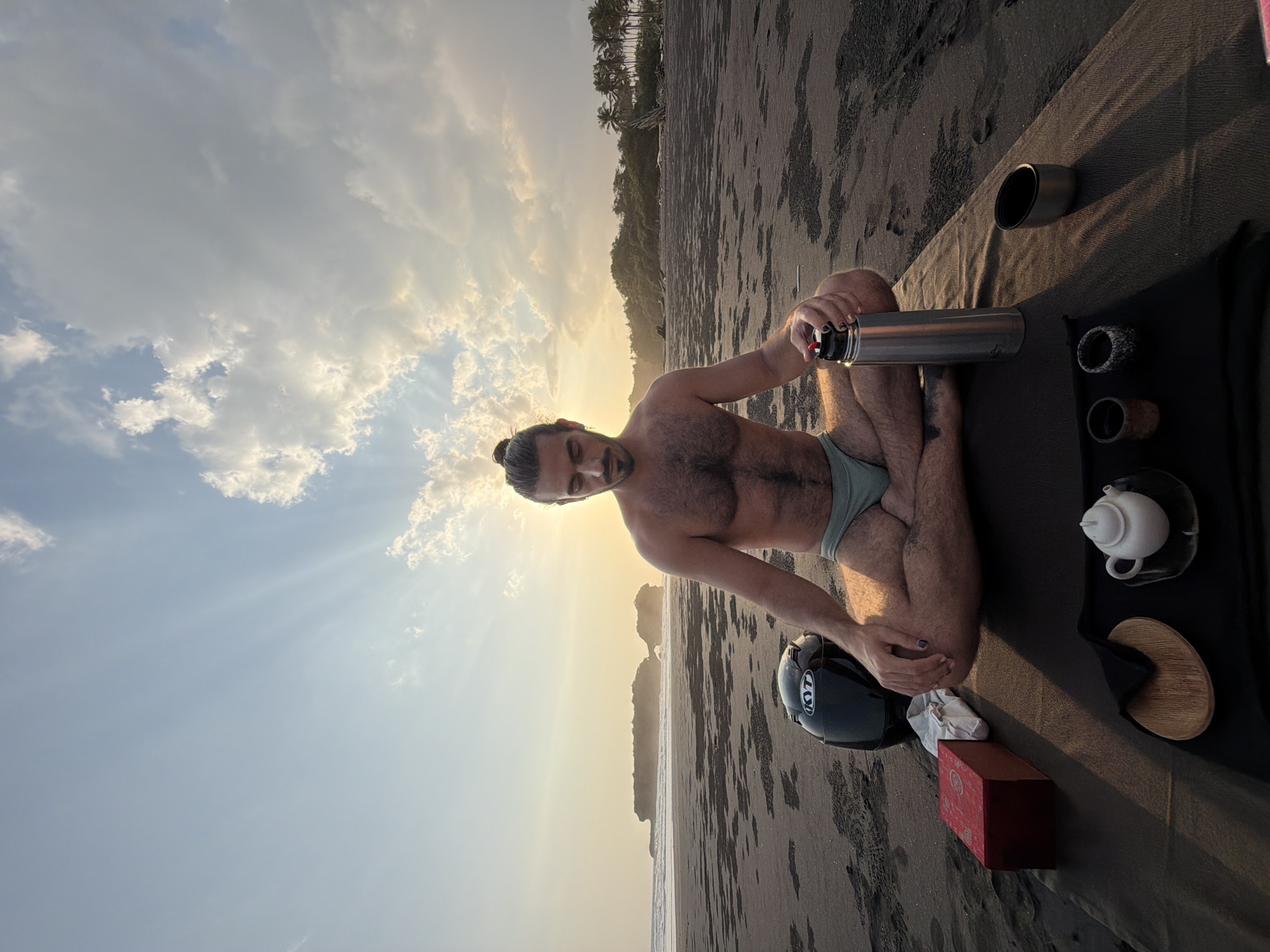 A man is meditating on a beach in Selemadeg, Indonesia, during sunset. He is sitting cross-legged on a dark mat, wearing light teal swimming briefs. His chest is bare, and he has dark hair pulled back into a bun. In front of him, on the mat, are various items for a tea ceremony: a red box, a white teapot, a wooden coaster, two small cups, and a metal thermos. To his left, a black motorcycle helmet rests on the sand. The beach is covered in dark volcanic sand, with ripples and footprints visible. In the background, the ocean meets the shore, and a rocky outcropping is visible in the distance. Lush green vegetation, including palm trees, lines the horizon. The sky is a gradient of soft blue and orange, with scattered clouds illuminated by the setting sun, casting long shadows across the sand.