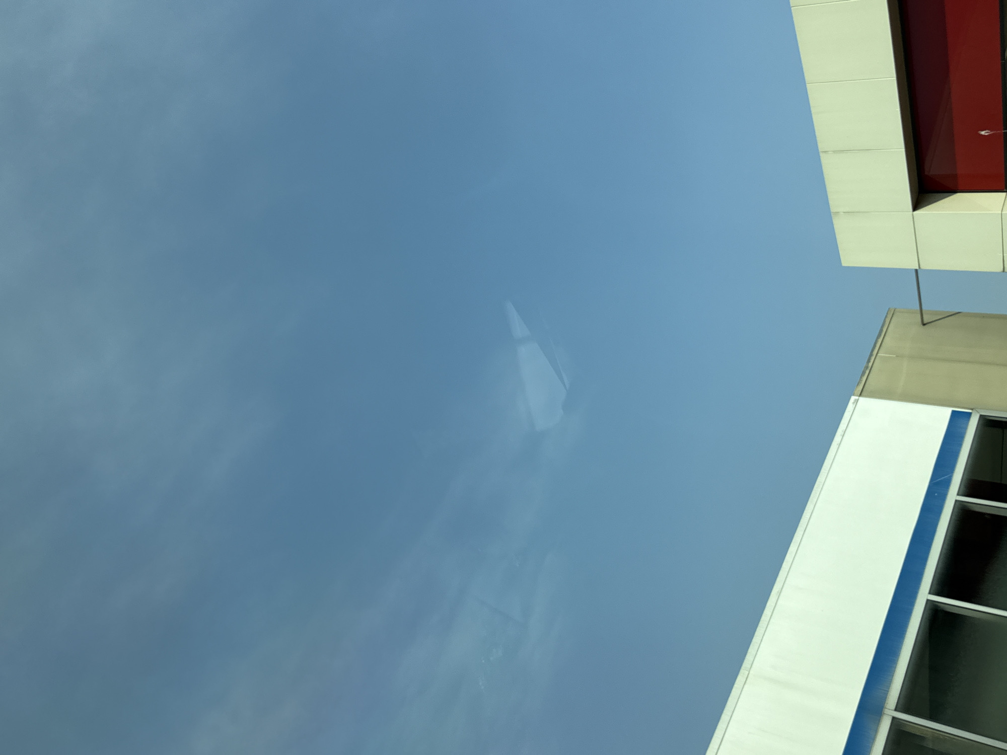 The image shows a clear blue sky with some faint, wispy clouds. Below the sky are the upper portions of two buildings. The building on the left is a long, white structure with a blue stripe running horizontally across its top. It has several large windows. The building on the right is a lighter colored structure with a red-colored section visible at the bottom. There are no people or animals visible in the image. The scene appears to be taken from a low angle, looking up at the sky and the buildings. The location is identified as Bad Sachsa, Germany.