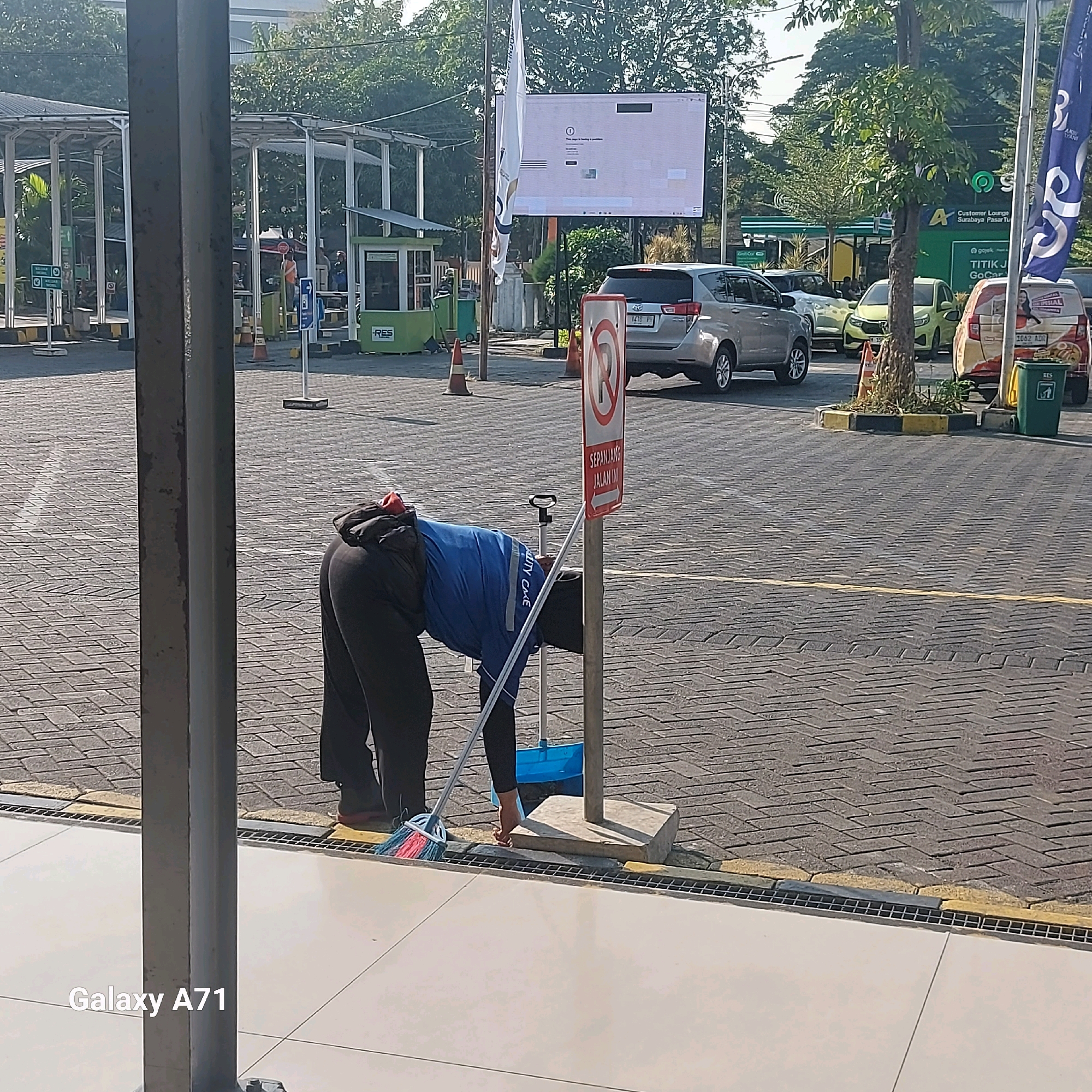 The image depicts an outdoor scene, likely a parking lot or public area in Surabaya, Indonesia. A person, wearing dark pants and a blue shirt with white lettering that appears to say "UTILITY CARE," is bent over and sweeping the paved ground with a broom. A blue dustpan is visible next to a metal pole with a sign attached. The sign is red and white, featuring a "P" with a diagonal line through it, indicating no parking, and text below that reads "SEPANJANG JALAN INI" which translates to "ALONG THIS ROAD." In the background, several cars are parked, including a silver SUV and a yellow car. A green building with "RES" and a logo that resembles "GoJek" is visible. Trees and awnings are also present. The ground is paved with interlocking bricks. The overall atmosphere suggests a sunny day in a functional urban environment.