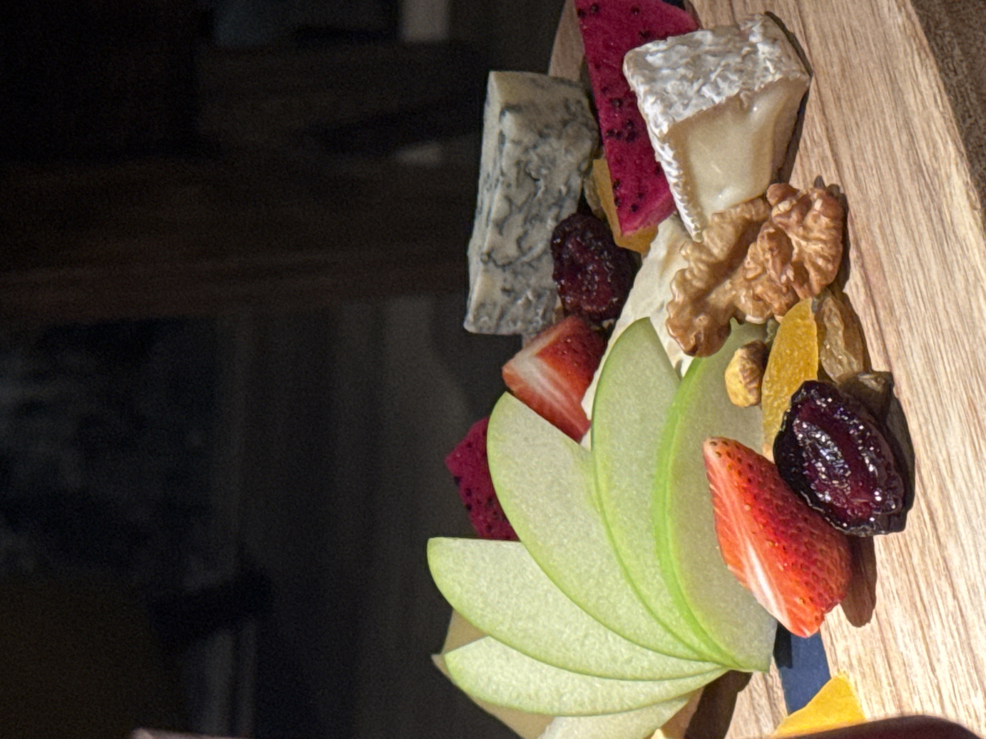 This media file captures a close-up of a cheese and fruit platter arranged on a wooden board. The platter features various cheeses, including a crumbly blue cheese and a soft white cheese with a rind. Accompanying the cheese are slices of vibrant pink dragon fruit, red strawberries, thinly sliced green apples arranged in a fanned pattern, dried fruit such as raisins and what appear to be dried apricots, and a whole walnut. A dark, glossy dried fruit, possibly a plum or cherry, is also visible. The overall presentation suggests a sophisticated snack or appetizer. The lighting is dramatic, with a dark background that contrasts with the brightly lit food items, creating a sense of depth and highlighting the textures and colors of the ingredients. The wooden board has a visible grain, adding to the rustic yet elegant aesthetic. The setting is likely indoors, given the controlled lighting and dark background. There is no visible text in the media file. The focus is entirely on the arrangement and appealing visual qualities of the food.