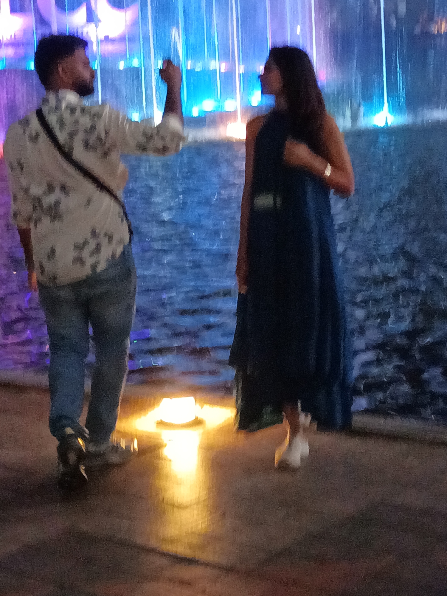 The image shows two people standing near a water fountain with colored lights. A man on the left, wearing a patterned shirt and jeans, is holding up a phone as if taking a picture. A woman on the right is wearing a dark blue dress and white shoes. The water fountain behind them has vertical streams of water illuminated by blue and purple lights. The ground is paved, and there is a bright yellow light reflecting on the ground in the foreground. The overall scene suggests a nighttime setting, possibly at a tourist attraction or public space. The location is identified as Kuala Lumpur, Malaysia.