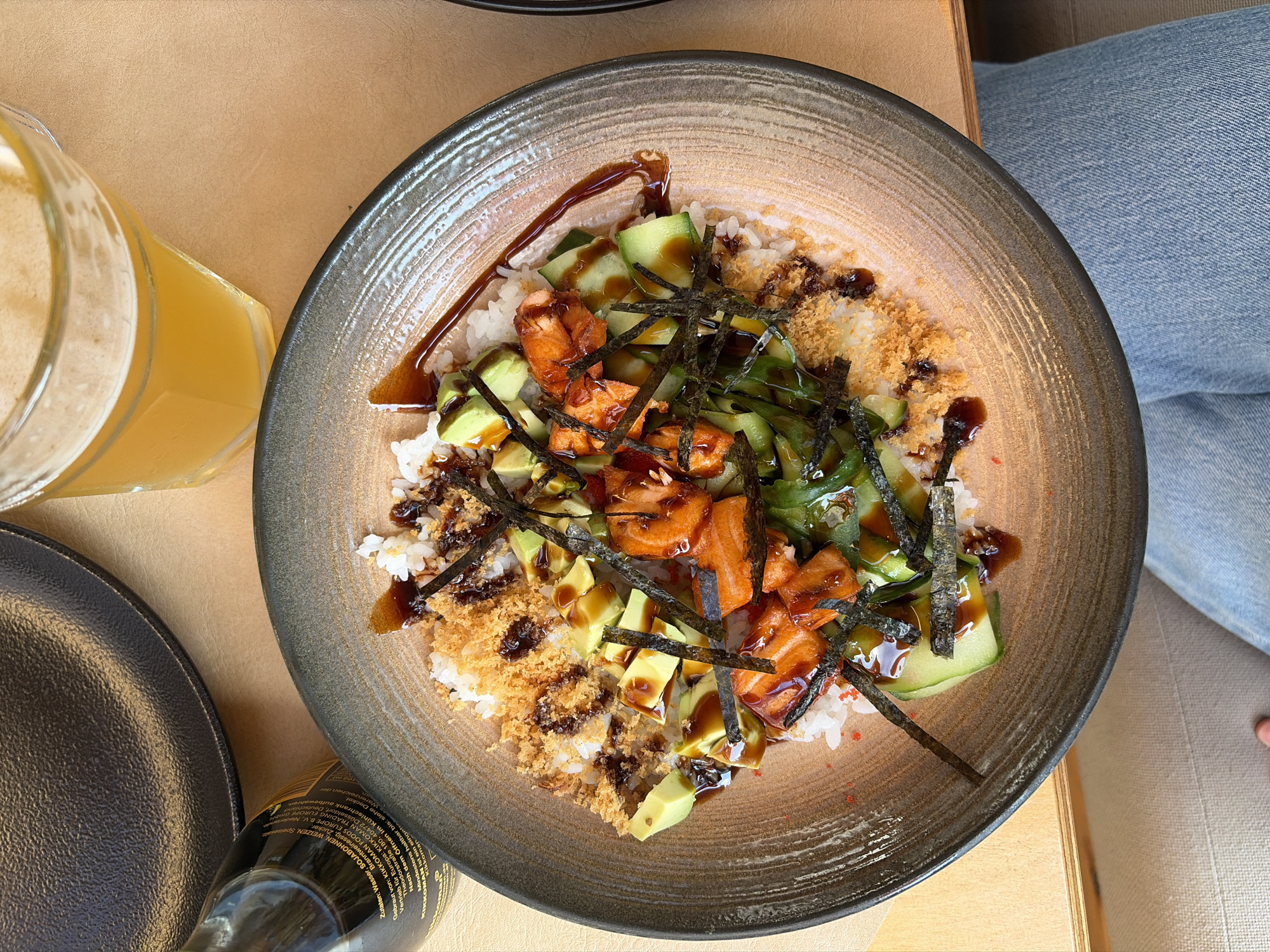 The image displays a flat lay composition centered around a large bowl of a deconstructed sushi or poke bowl dish, presented on a light brown table surface. The bowl, round with an earthy-toned, textured rim, contains white rice topped with cubed salmon, sliced avocado, thinly sliced cucumber, and strips of dark green nori (seaweed). A dark brown sauce, likely soy-based, is drizzled over the ingredients and pools at various points. Portions of the dish are also covered with a golden-brown, crispy topping, resembling panko breadcrumbs or tempura flakes. Small red specks, possibly masago or tobiko, are visible among the ingredients.
To the upper left of the main bowl, a dark glass Kikkoman brand soy sauce bottle is partially visible, showing German text on its label. Above the soy sauce bottle, a segment of an empty, dark gray or black textured plate is present. To the upper right, a clear glass contains an amber liquid with a foam head, consistent with beer or a similar carbonated beverage.
The lower portion of the image shows the thighs and knees of a person wearing light blue denim jeans, indicating a first-person or close-up perspective of someone seated at the table. A small portion of what appears to be a chair or bench cushion is visible below the table edge on the left. The lighting is bright and even, casting soft shadows. The specific city and location are not identifiable from the image.