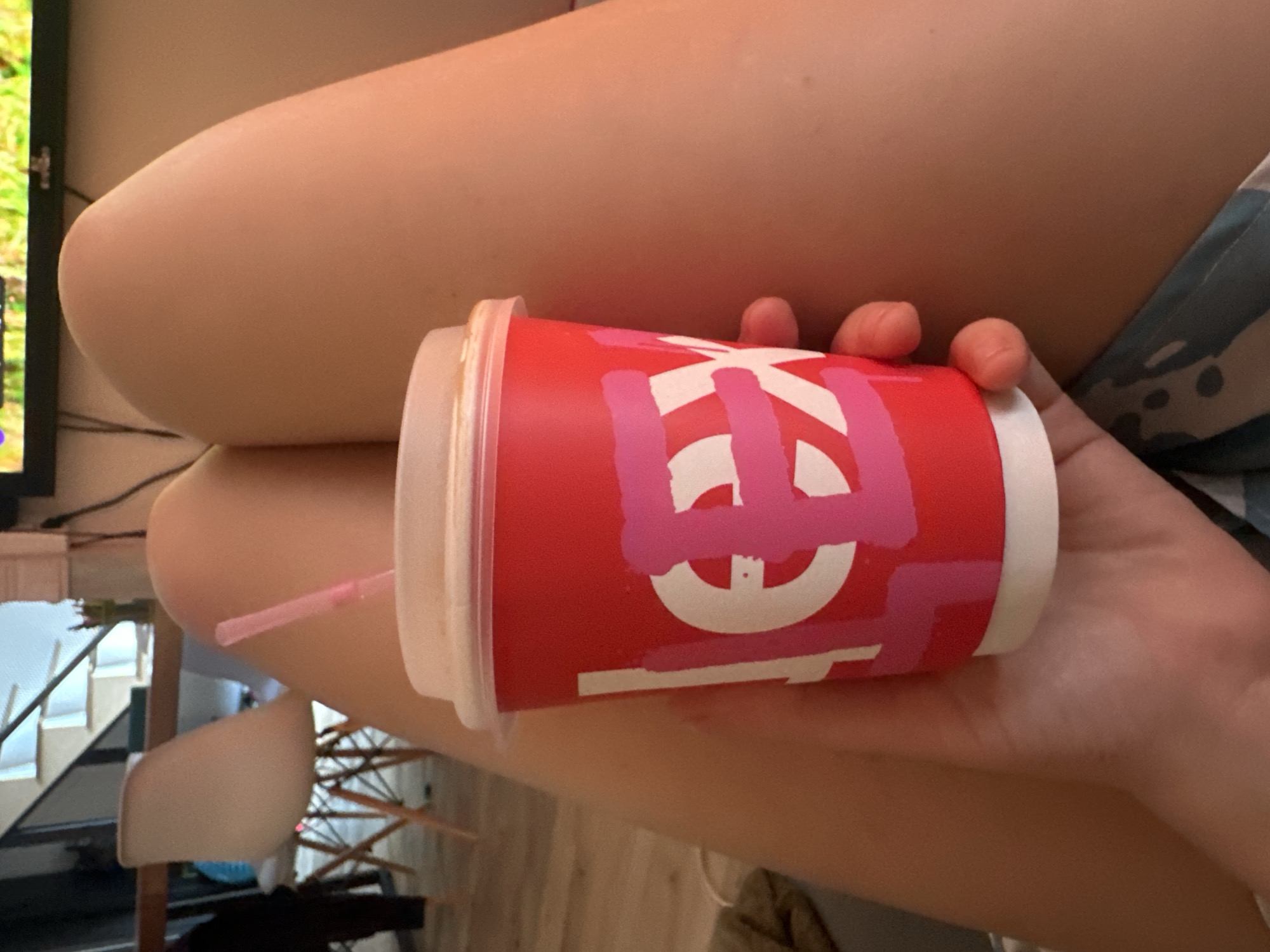 A person is holding a red disposable cup with a white lid and a pink straw. The cup has a white graphic design with pink brush strokes over it. The person's legs are visible, one of which is resting on a patterned surface. In the background, a chair and stairs are partially visible. The overall scene suggests an indoor setting, possibly a home.