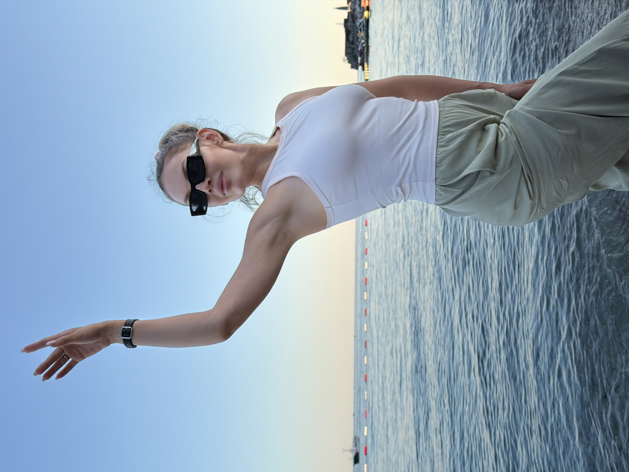 A woman, with blonde hair pulled back, wears black sunglasses and a white tank top. She is posing by the sea, with her left arm extended and her hand open. She is wearing a black watch on her left wrist and a ring on her left ring finger. She is also wearing loose, light-colored pants. The sea is calm with some ripples, and a line of buoys floats on the water. In the background, a city skyline with buildings and lights can be seen on the horizon. The sky is clear and a pale blue, suggesting it is either early morning or late evening. The overall mood is relaxed and serene. The image was taken in Budva, Montenegro.