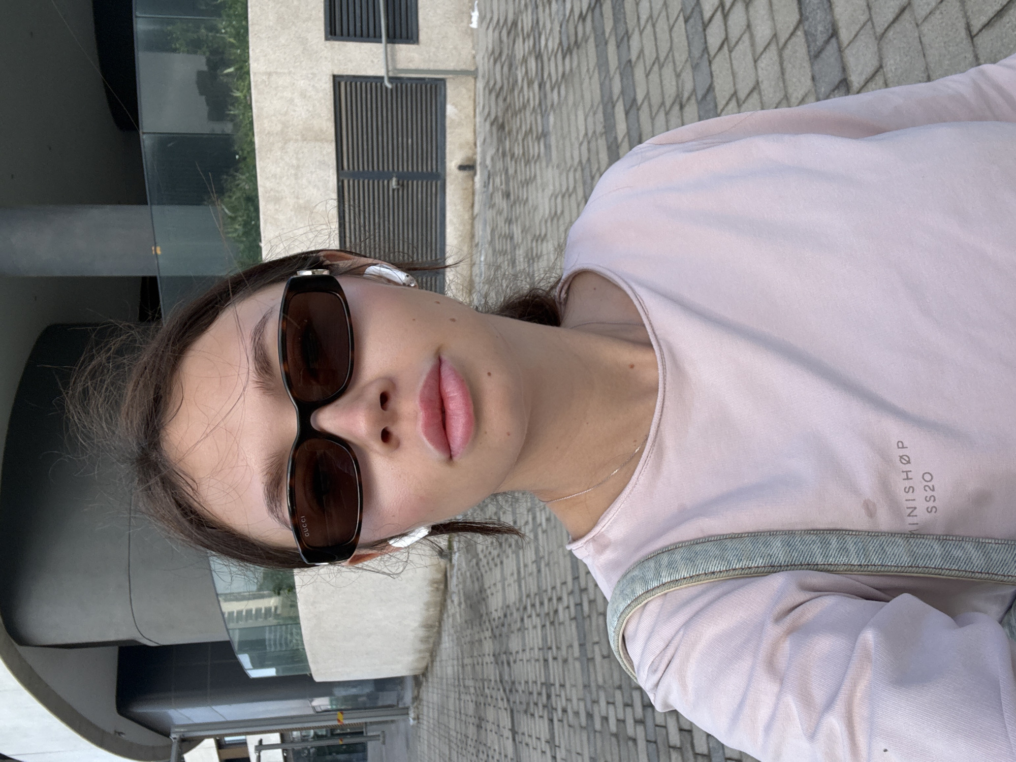 A young woman is pictured in a selfie-style image taken outdoors. She has brown hair, light skin, and is wearing dark sunglasses, one of which has the word "Gucci" visible on the side. She also has white earbuds in her ears. She's wearing a light pink top with a denim-trimmed neckline, with lettering "MINISHØP SS20" on the shirt. The background includes modern architecture with glass windows and gray tiled pavement. Based on the architectural style and background, it is probable that the location is in Tel Aviv, Israel.