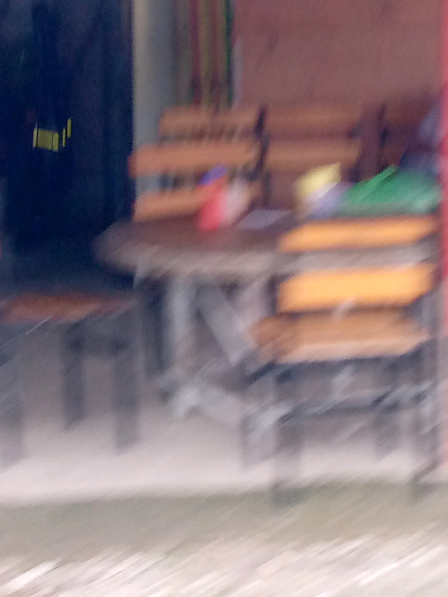 The image displays a blurred scene of an outdoor seating area, likely a cafe or restaurant, with empty wooden chairs and tables. The setting appears to be in Glokaron, Indonesia, as indicated by the presence of a dark blue facade with faint yellow markings that may be a business sign. The background is a soft, indistinct wall with vertical lines of various colors, possibly indicating a mural or decor. The lighting is dim, suggesting it might be dusk or an overcast day. There are no people or active interactions visible in the frame.