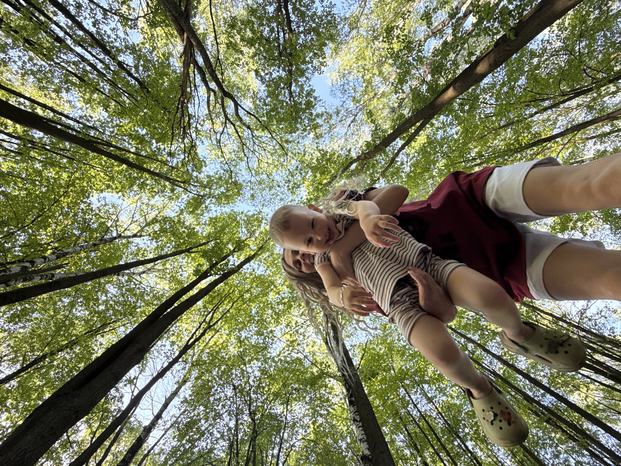 A low-angle shot shows a woman holding a baby aloft against a backdrop of tall trees with green leaves, seen from below, in what appears to be a forest. The sky is visible through the canopy. The woman wears a maroon top and white shorts and olive-green Crocs. The baby is wearing a striped outfit and olive-green Crocs. The location is a forest in the city of Шебунино, Russia.