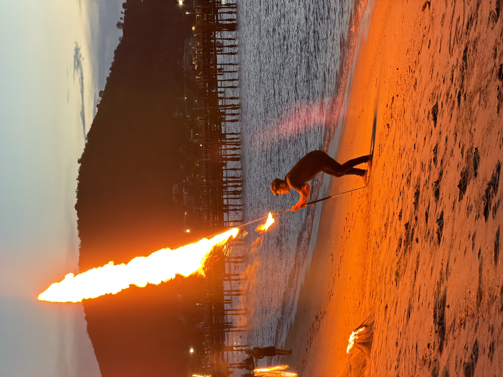The image depicts a nighttime scene on a beach in Koh Samui, Thailand, likely during a fire performance. A man is standing on the sand, holding a pole with a large flame erupting from its end, reaching high into the sky. To his left, a smaller fire burns on the beach. In the background, a wooden pier extends into the ocean, with lights illuminating structures on it and along the coastline. A silhouetted figure of a woman is visible on the beach near the water's edge. The ocean is calm, and the sky is a gradient of twilight colors. The sand is illuminated by the warm glow of the fires.